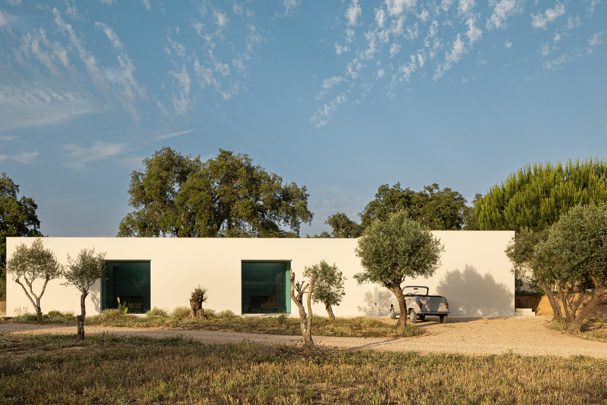 White stucco facade with dark window openings and olive trees along a gravel drive under cirrus clouds