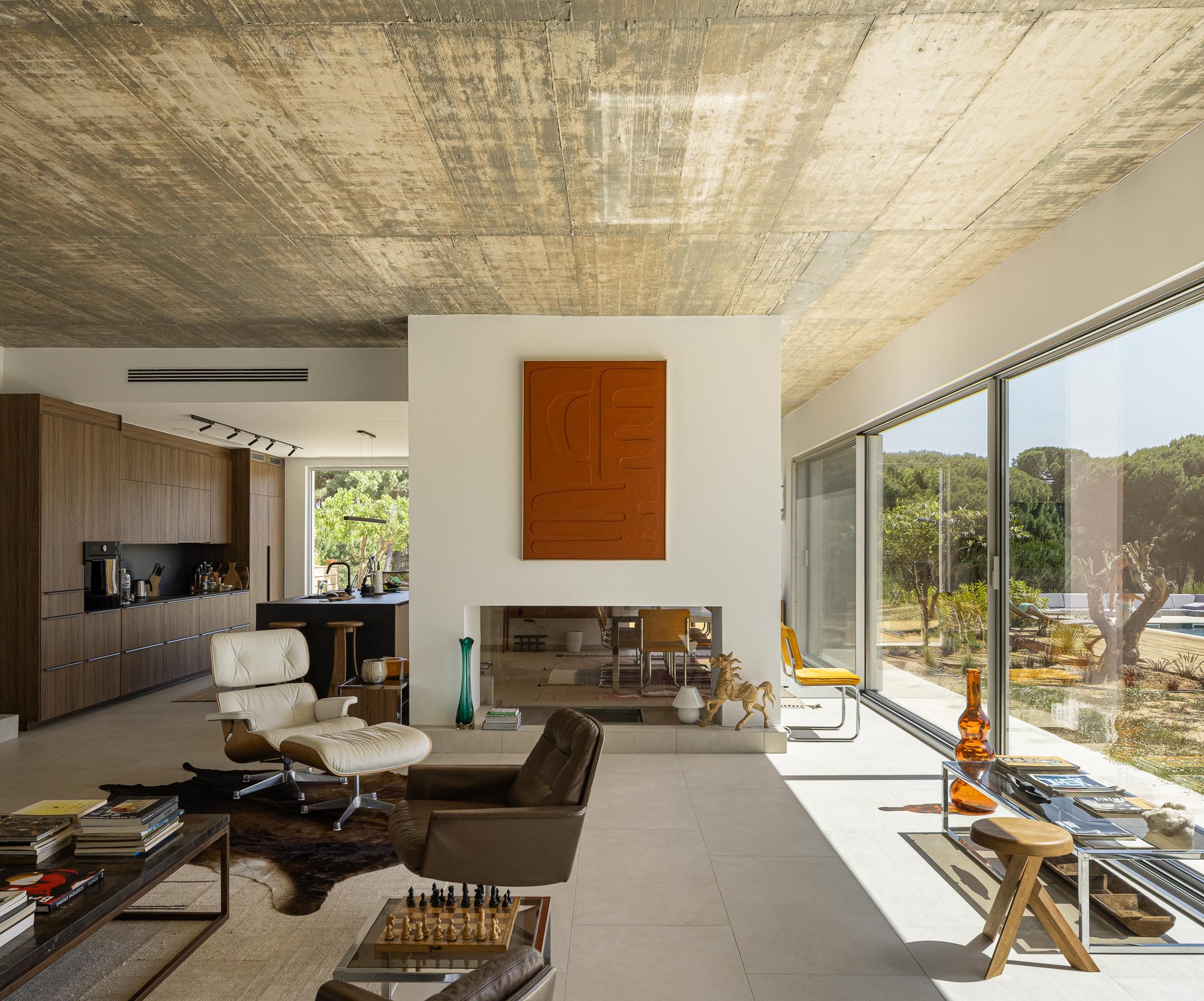 Living space with timber-lined ceiling and full-height glazing overlooking olive groves and a pool