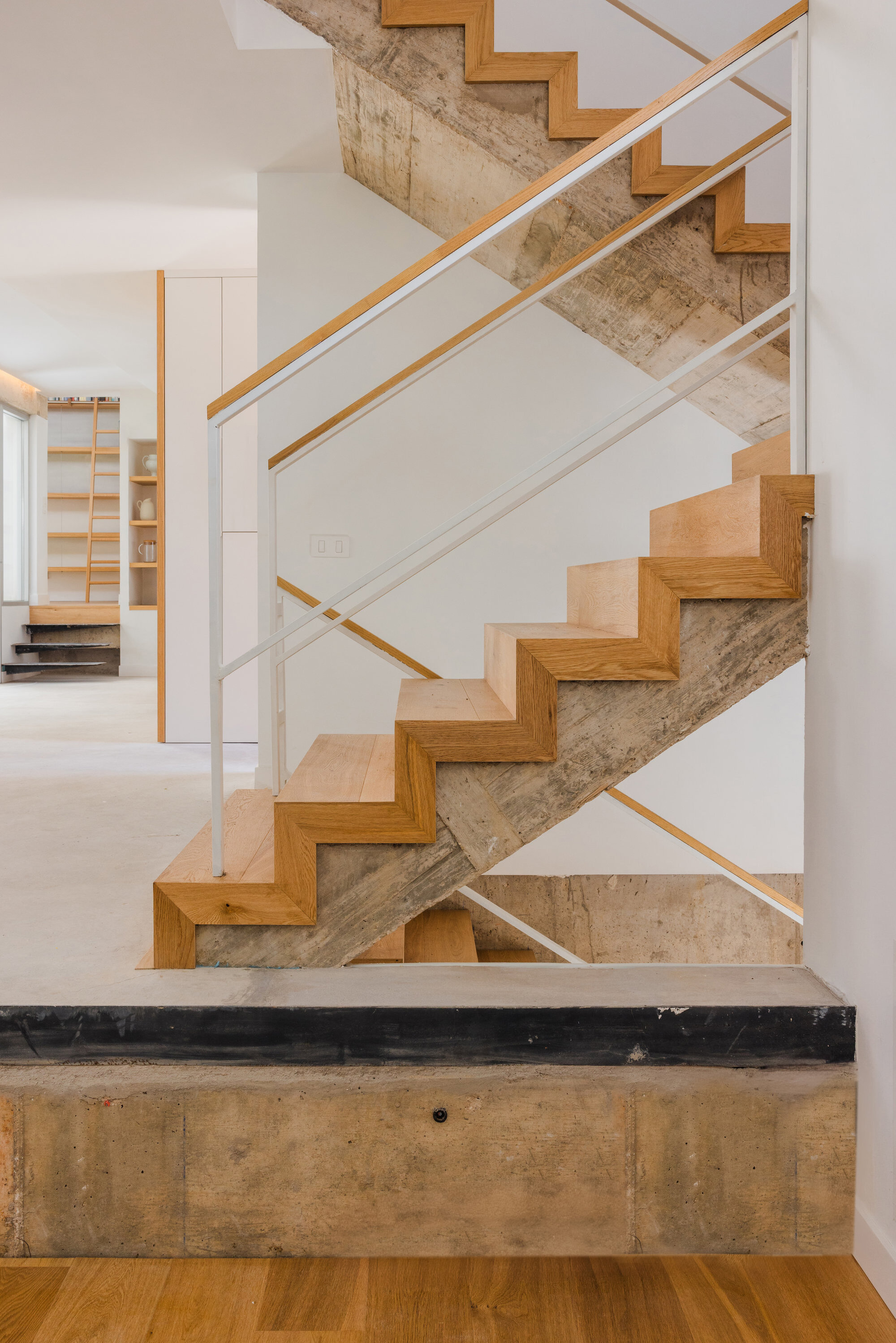 Oak staircase wrapping around exposed concrete core with white powder-coated steel handrails