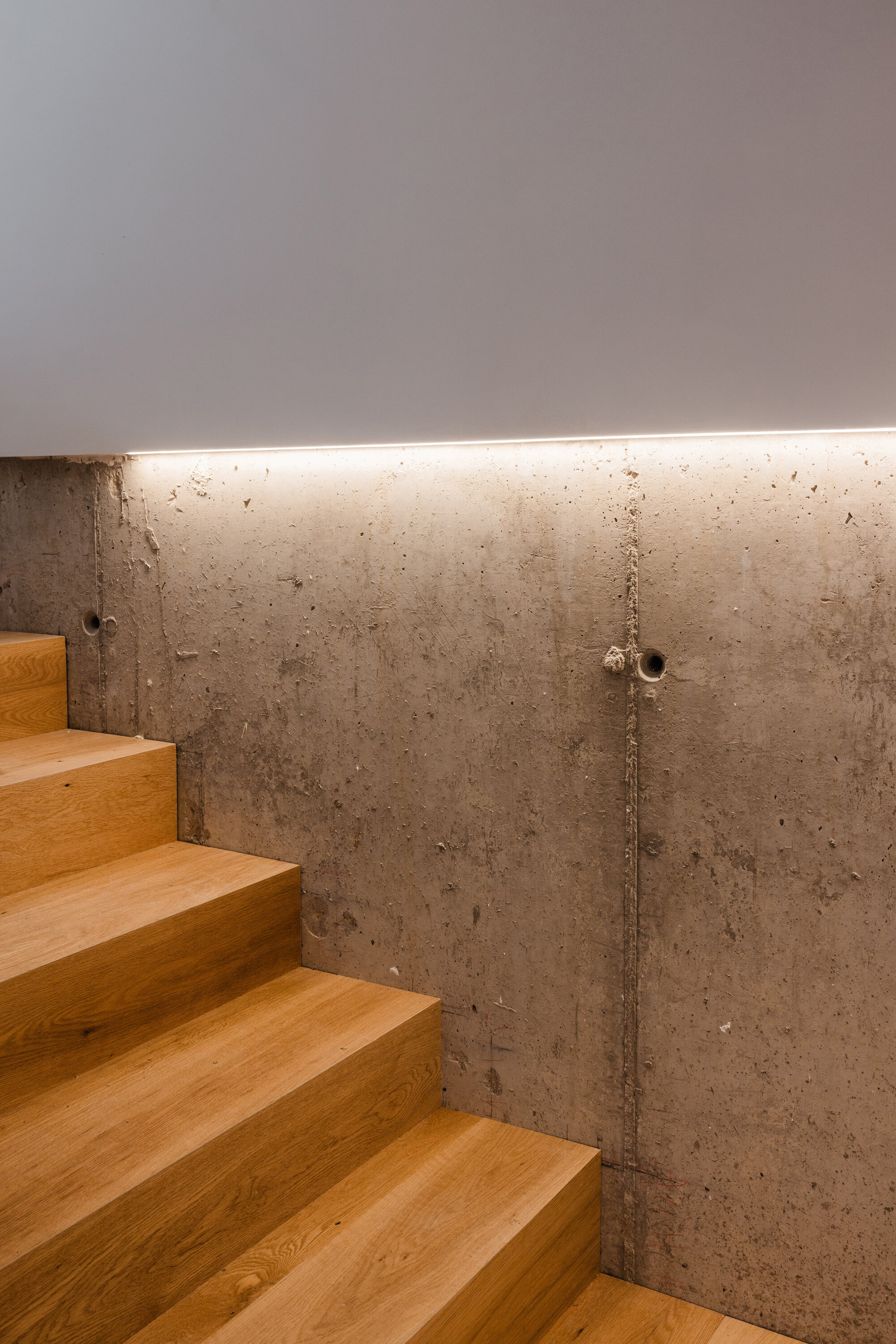 Oak stair descending along board-formed concrete wall with recessed linear lighting at ceiling