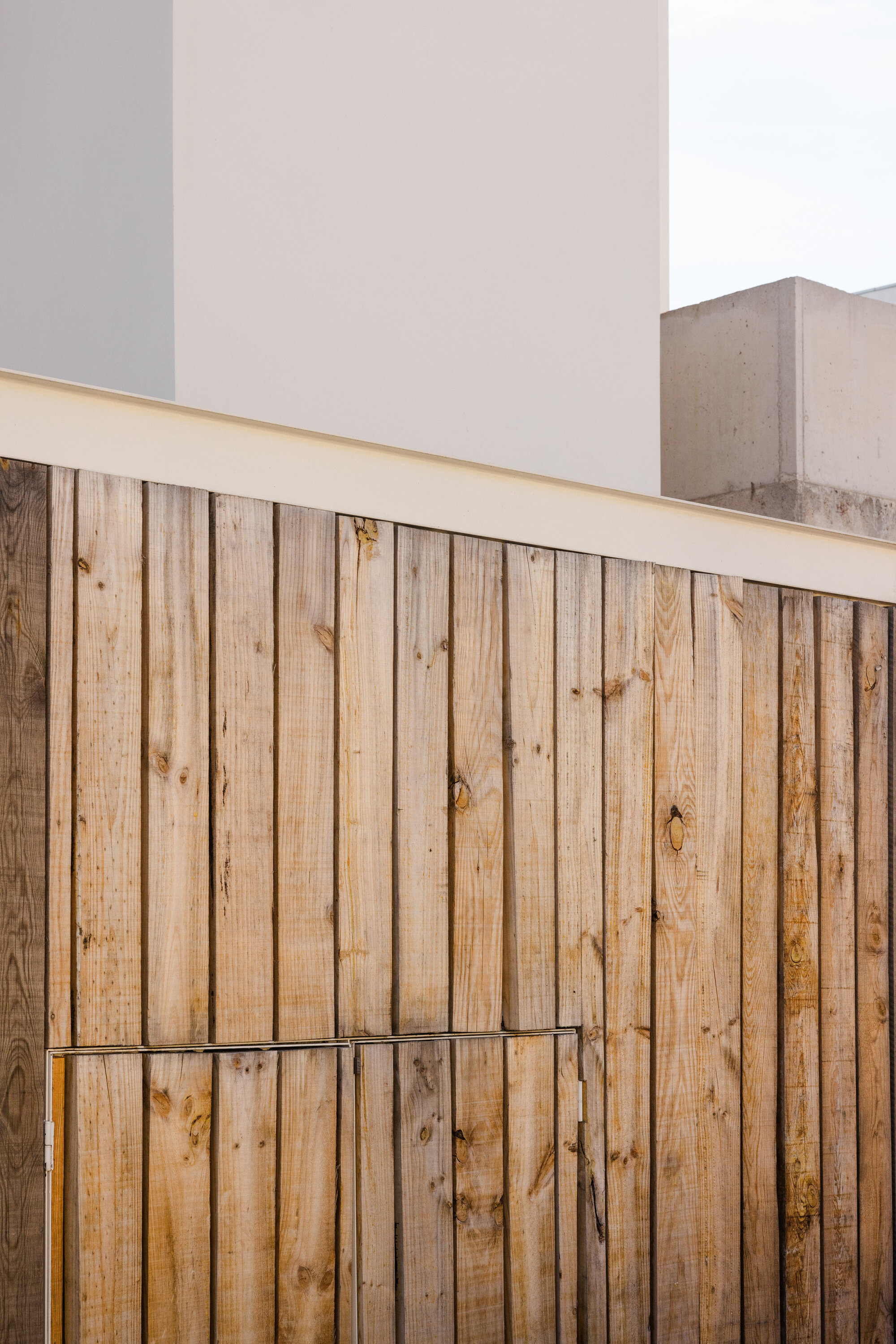Vertical timber slat fence beneath white horizontal coping against concrete wall and cloudy sky