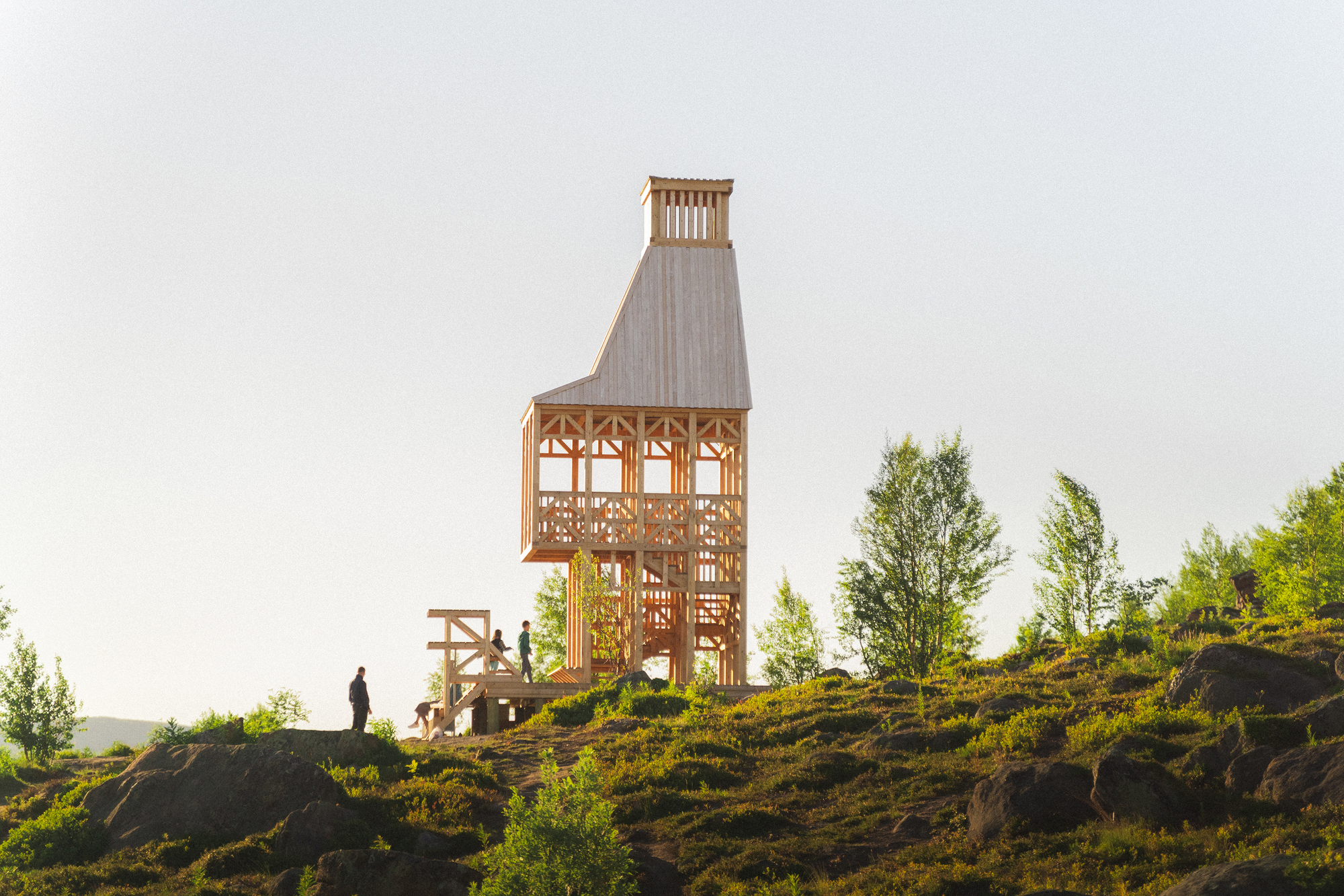 Timber observation tower with open lattice walls on a rocky hillside with visitors approaching at dusk