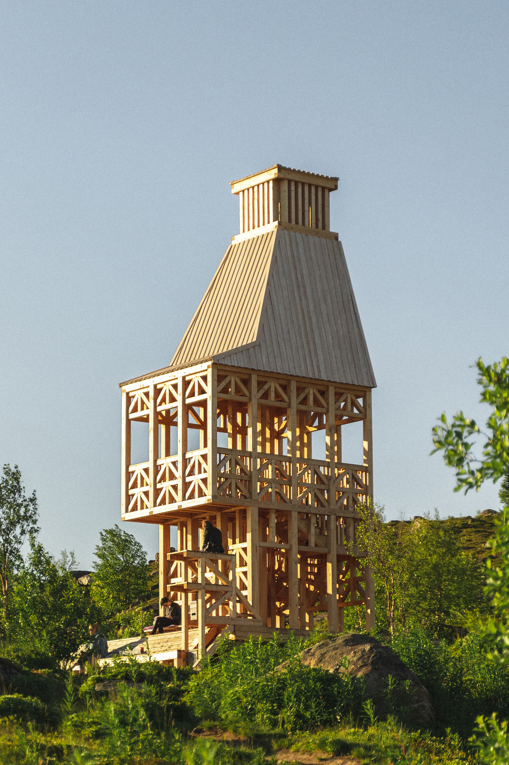 Freestanding wooden tower with tapered form and slatted cladding among birch trees in evening light