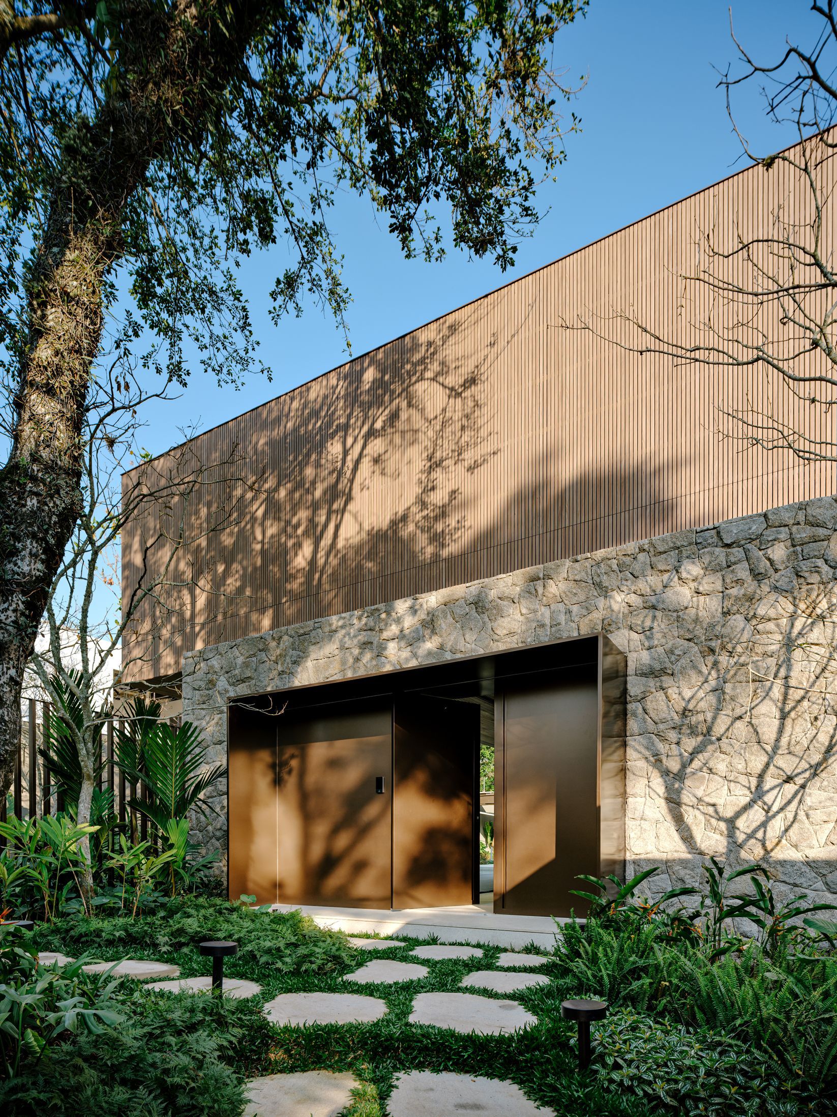 Stone base and vertical timber cladding facade with bronze entry doors framed by stepping stone path
