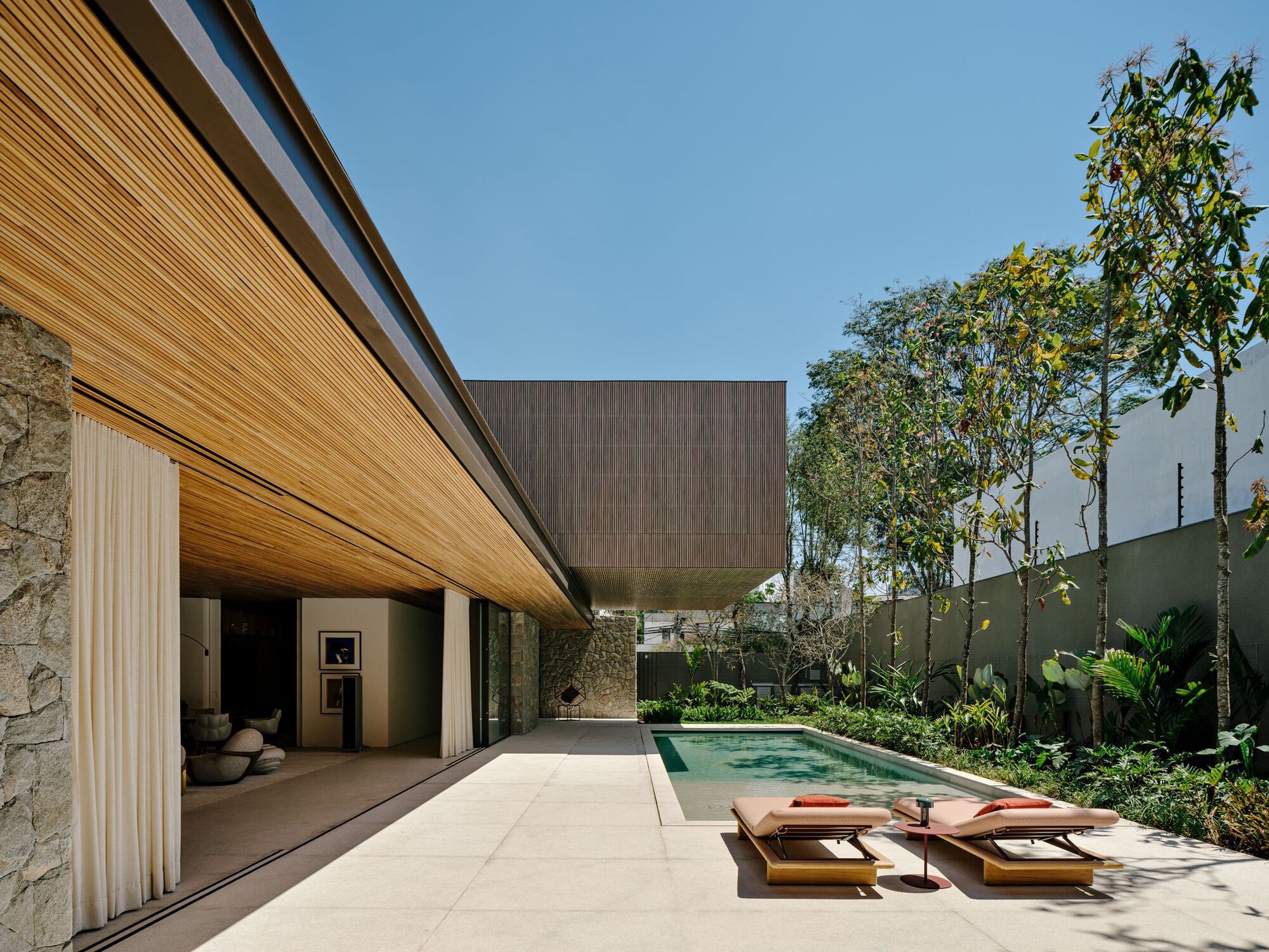 Pool terrace with timber-clad volumes and dark upper volume framed by tall trees under blue sky