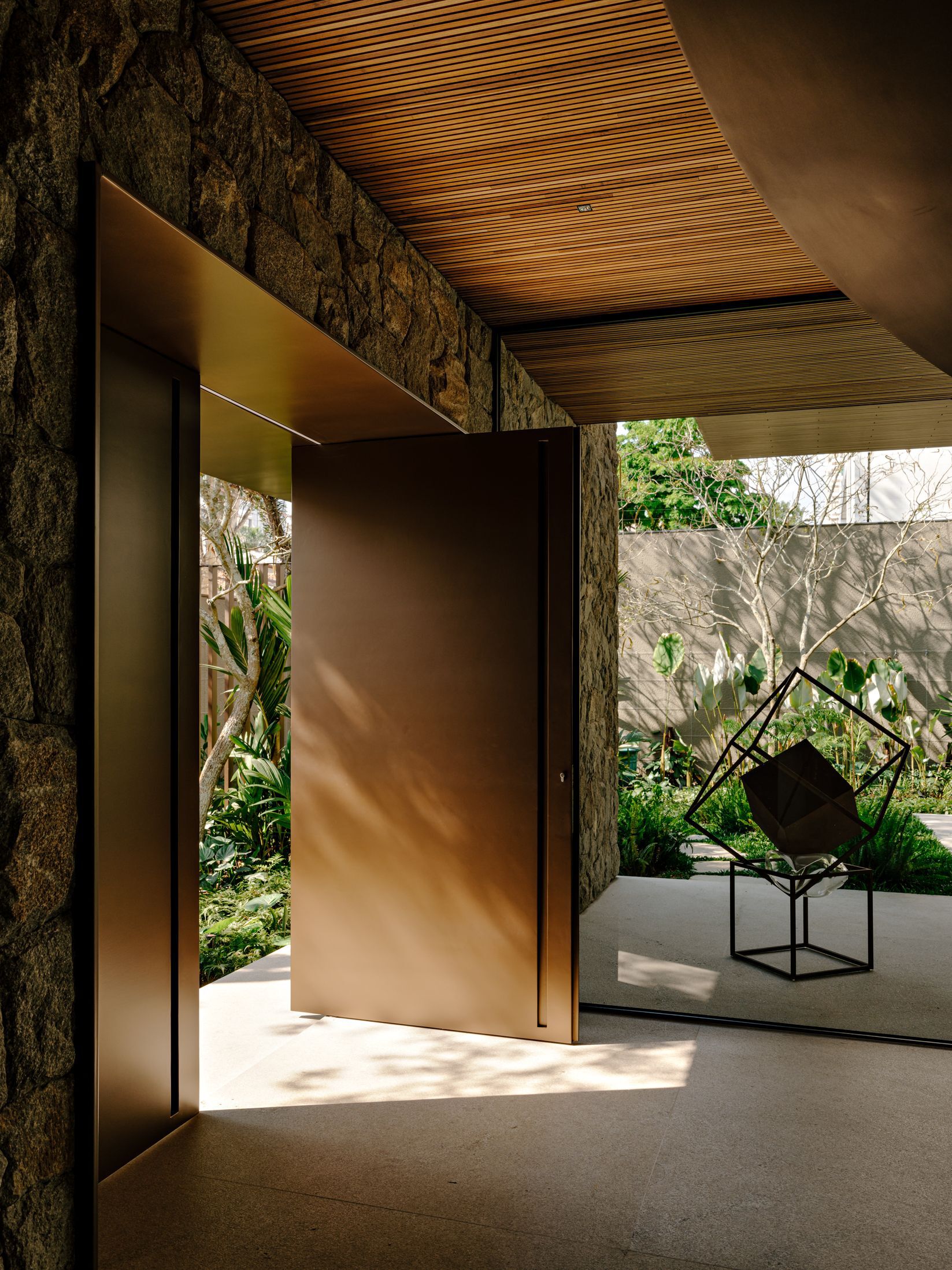 Pivoting bronze entry door opening to a garden courtyard with sculpture under a timber soffit