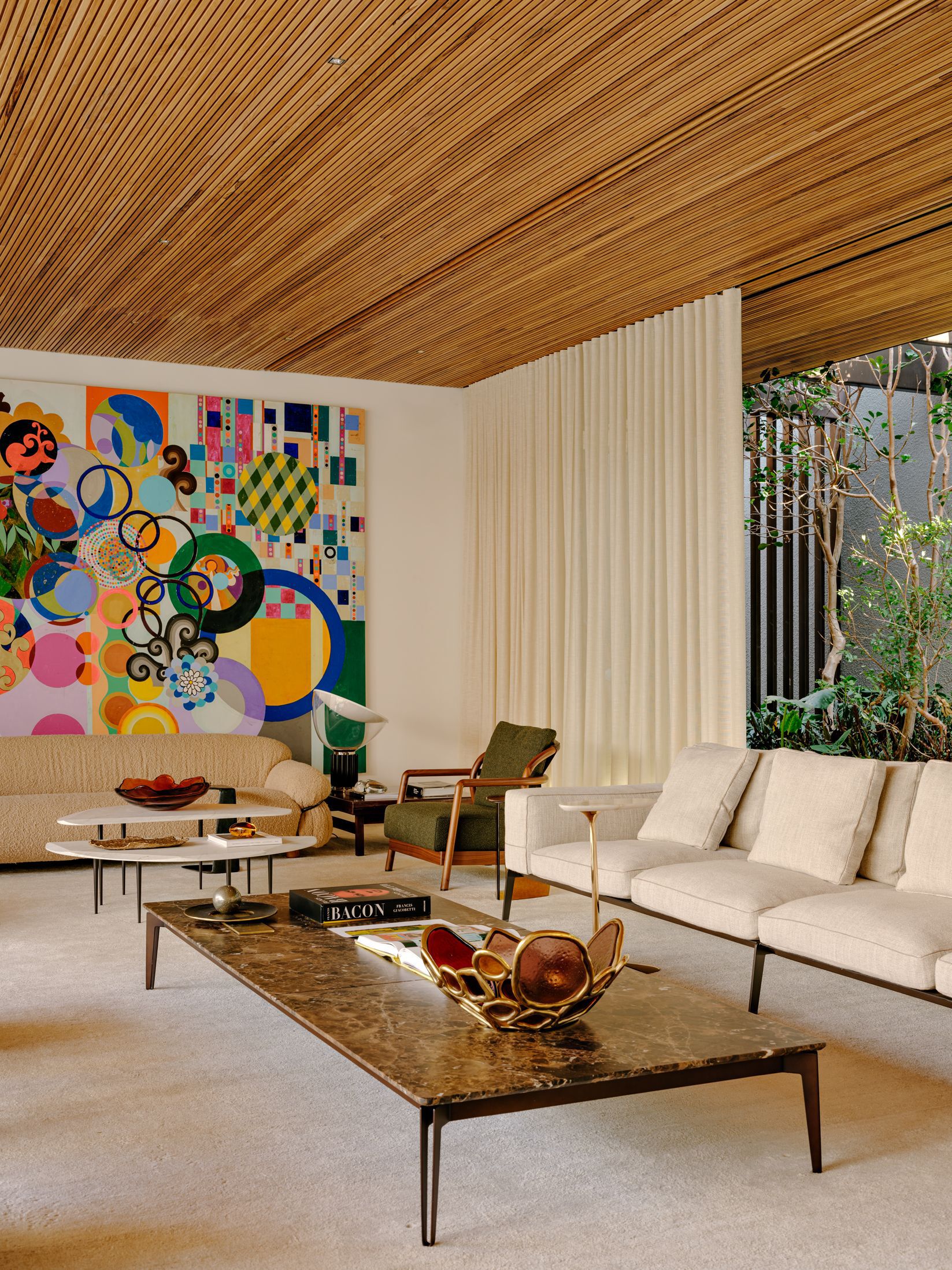 Living room view showing slatted timber ceiling, abstract mural, and glass wall to planted courtyard