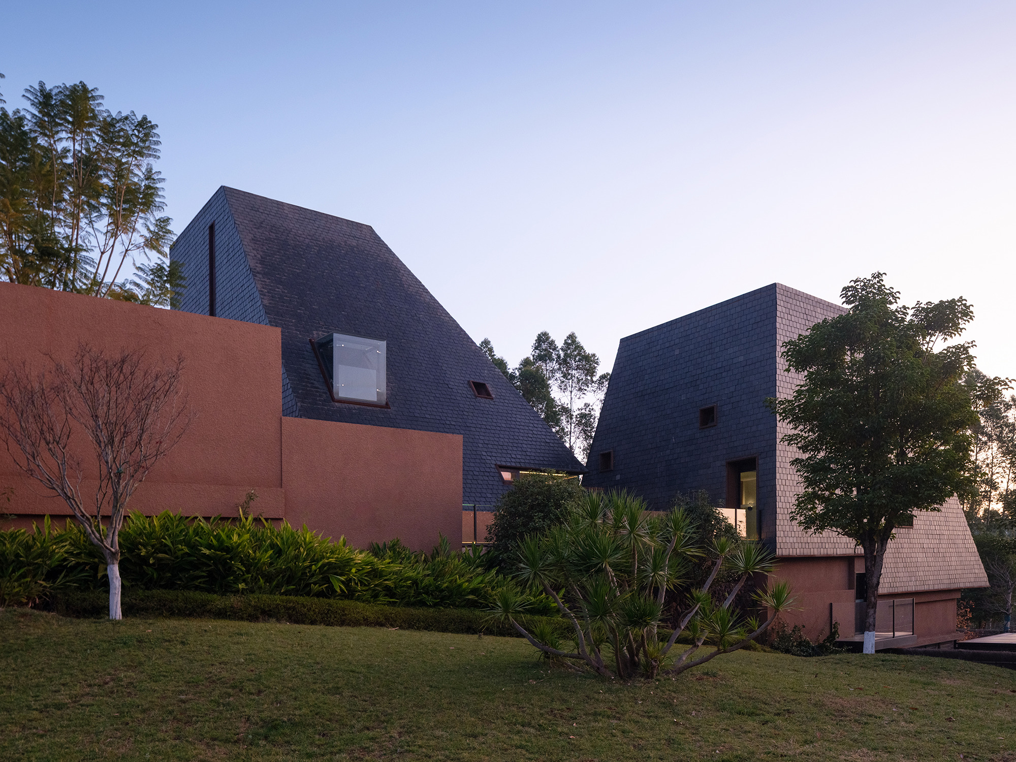 Clustered volumes clad in dark shingles rising above planted lawn and terracotta walls at dusk