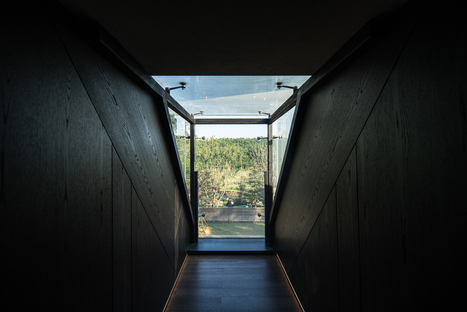 Dark timber-lined corridor with angled walls opening to glass door and landscape beyond
