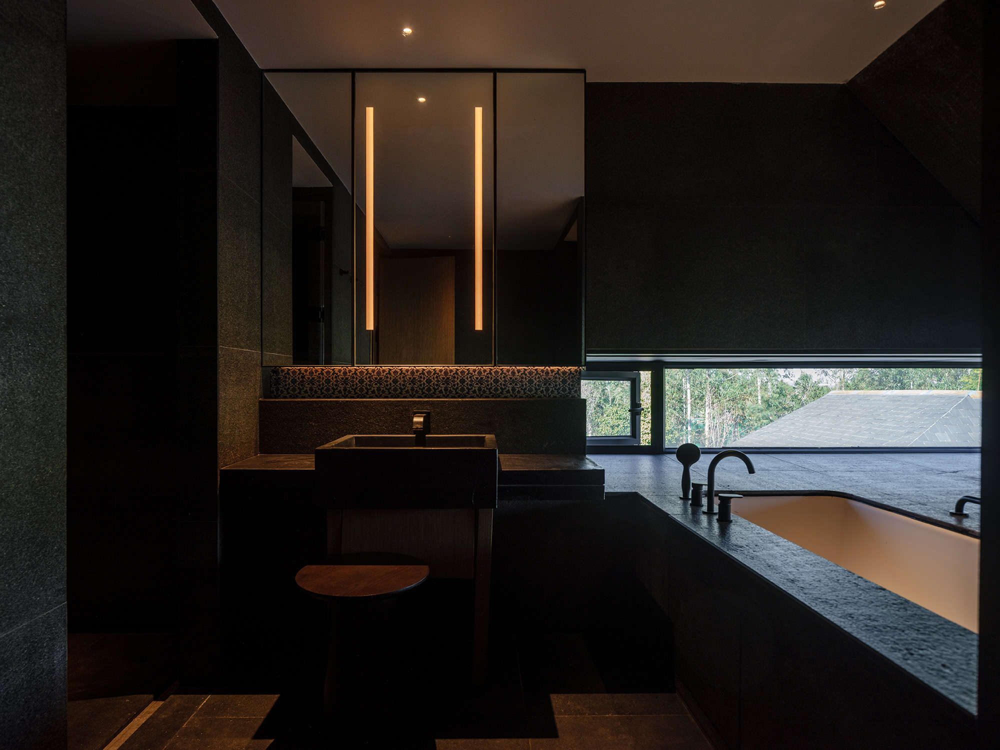 Dark bathroom vanity with vertical strip lighting above twin sinks and horizontal window overlooking treetops