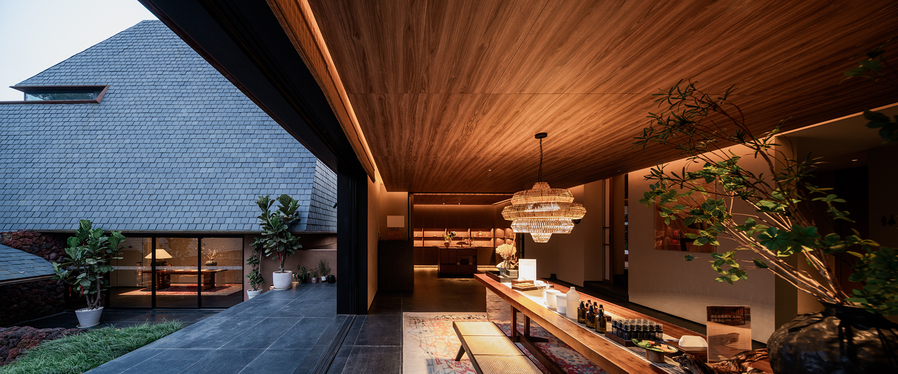 Covered outdoor terrace with timber ceiling opening to courtyard with tiled roof volume at dusk