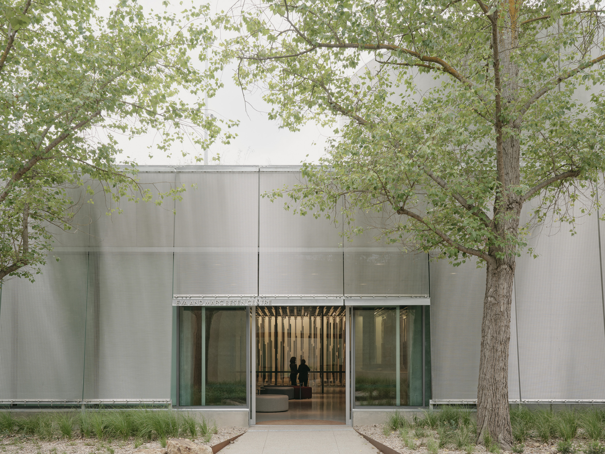 Entrance facade with translucent channel glass panels and glazed doors framed by existing trees and gravel beds