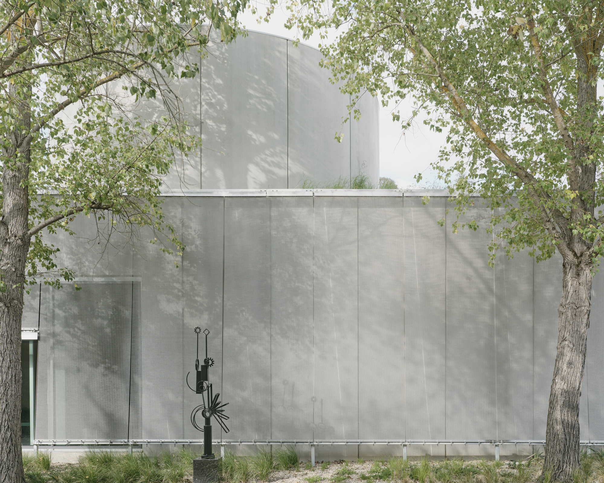 Textured concrete wall with vertical paneling beneath a protruding upper volume visible through dappled tree shadows