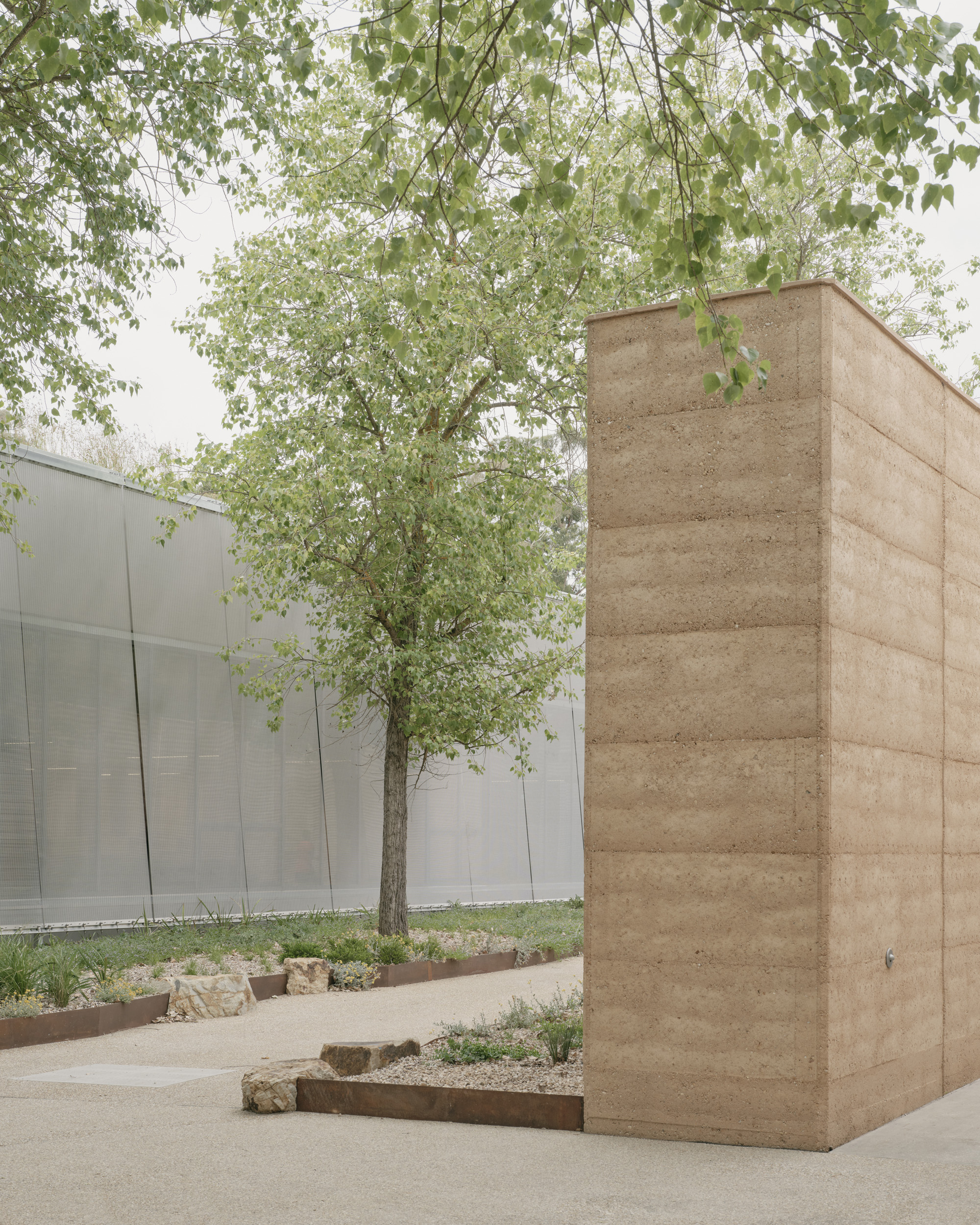 Rammed earth column beside a planted bed with tree and white metal panel facade beyond