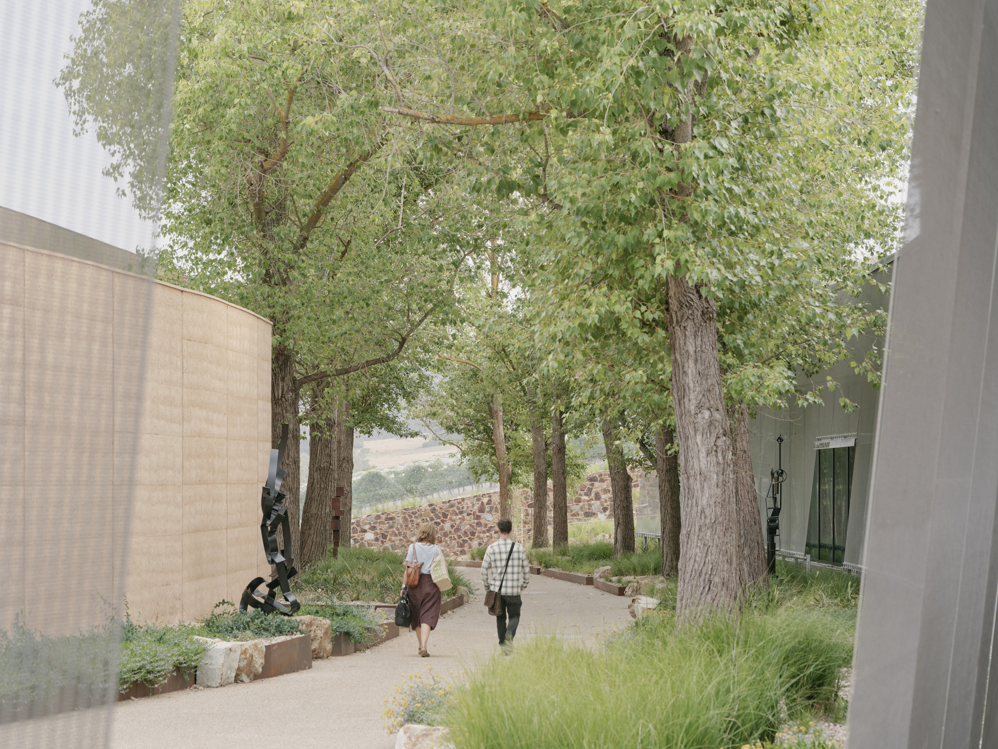Tree-lined pathway with visitors walking between rammed earth walls and stone edged planting beds