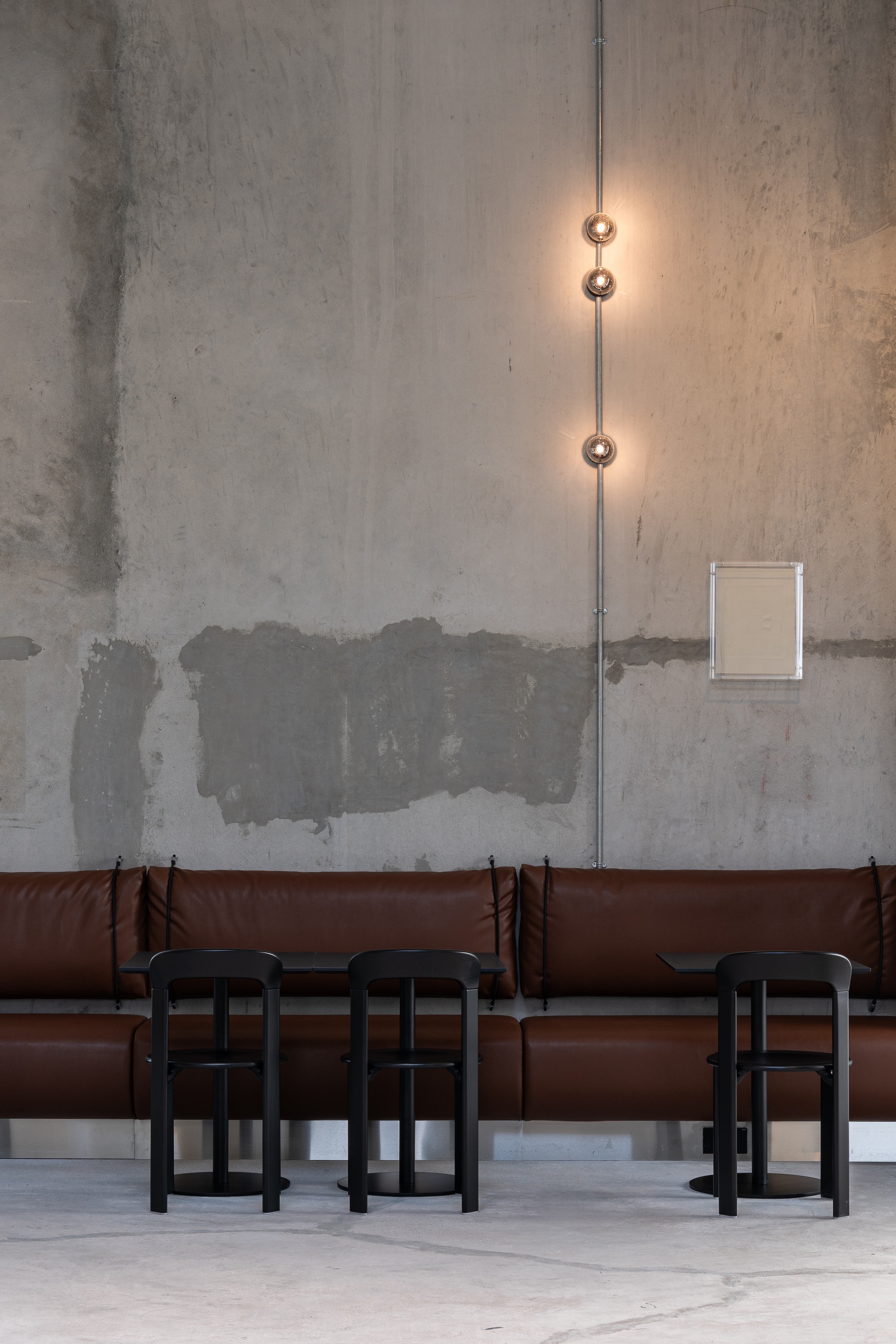 Brown leather banquette with black timber stools beneath a vertical pendant light on an unfinished concrete wall