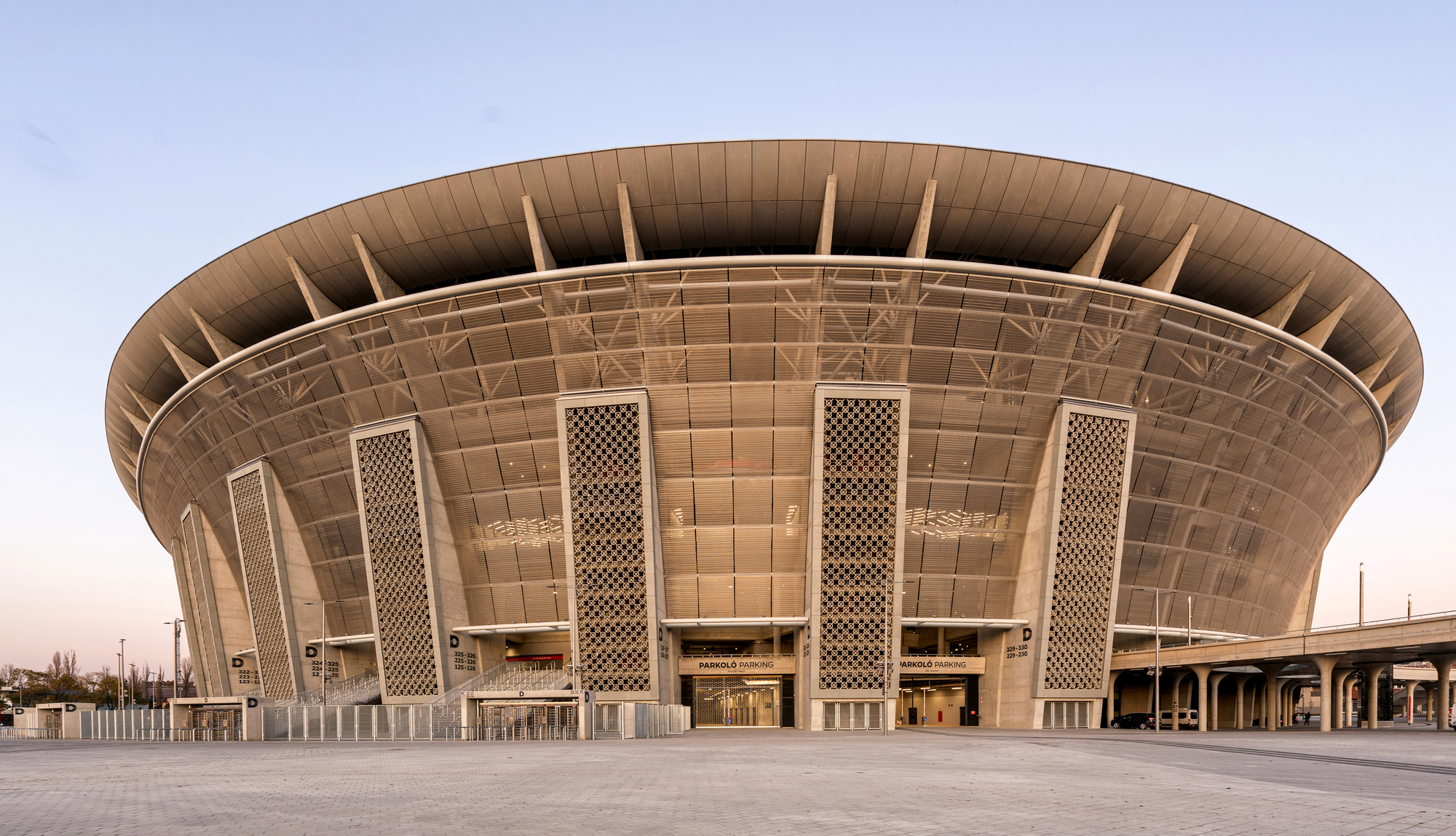 Circular facade with tiled panels and tall perforated screens under an overhanging roof at dusk