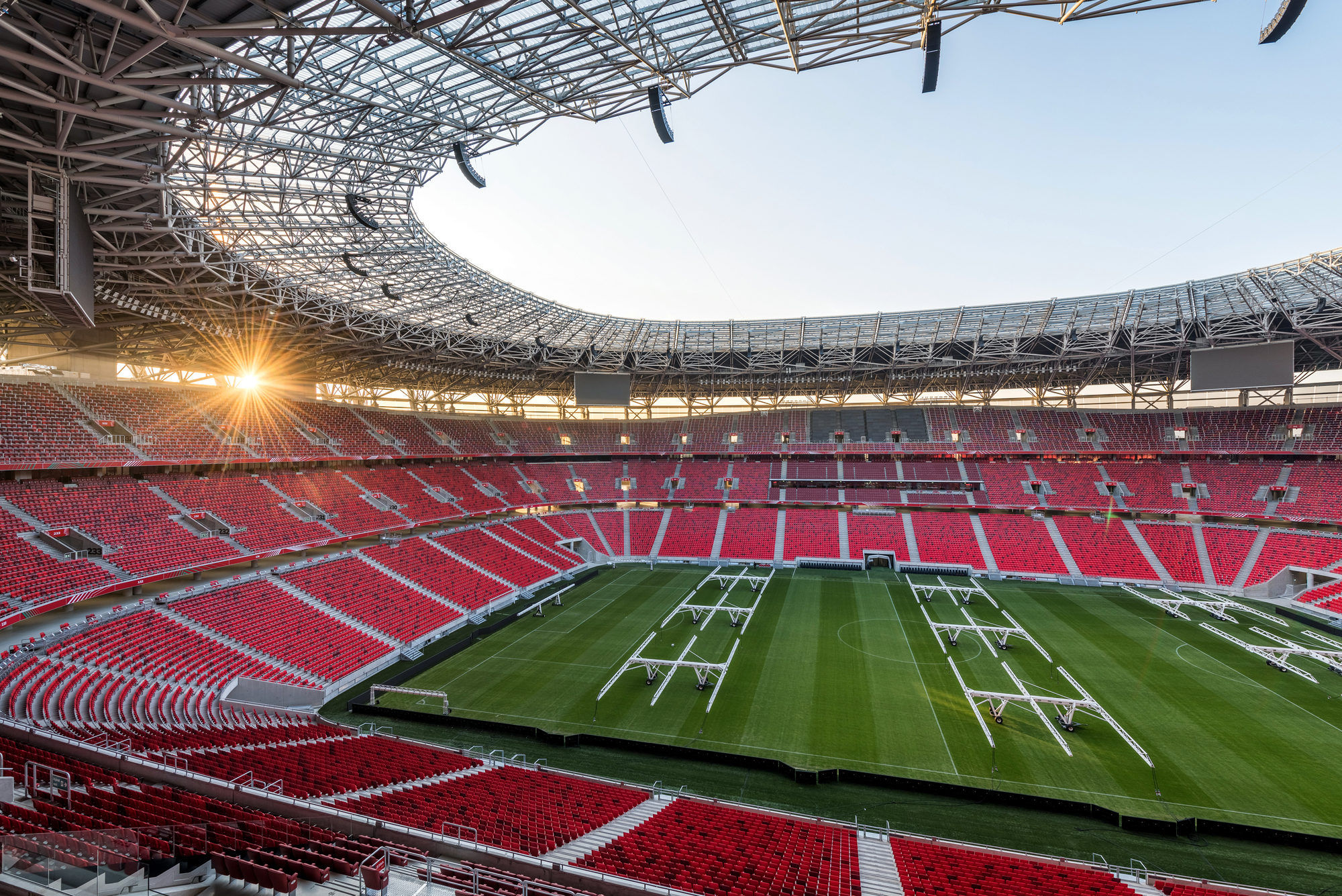 Stadium bowl with red seating tiers surrounding a green pitch under an open steel roof