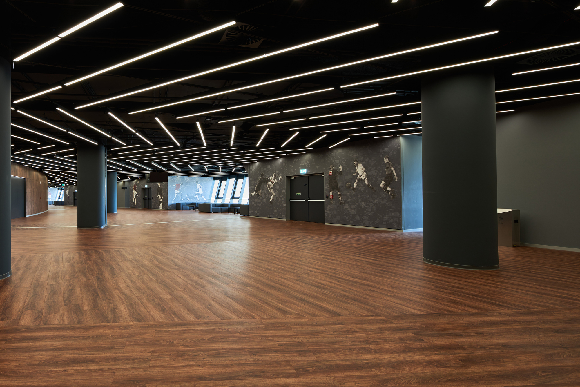 Interior lobby space with herringbone wood flooring and linear ceiling lights over dark columns and walls