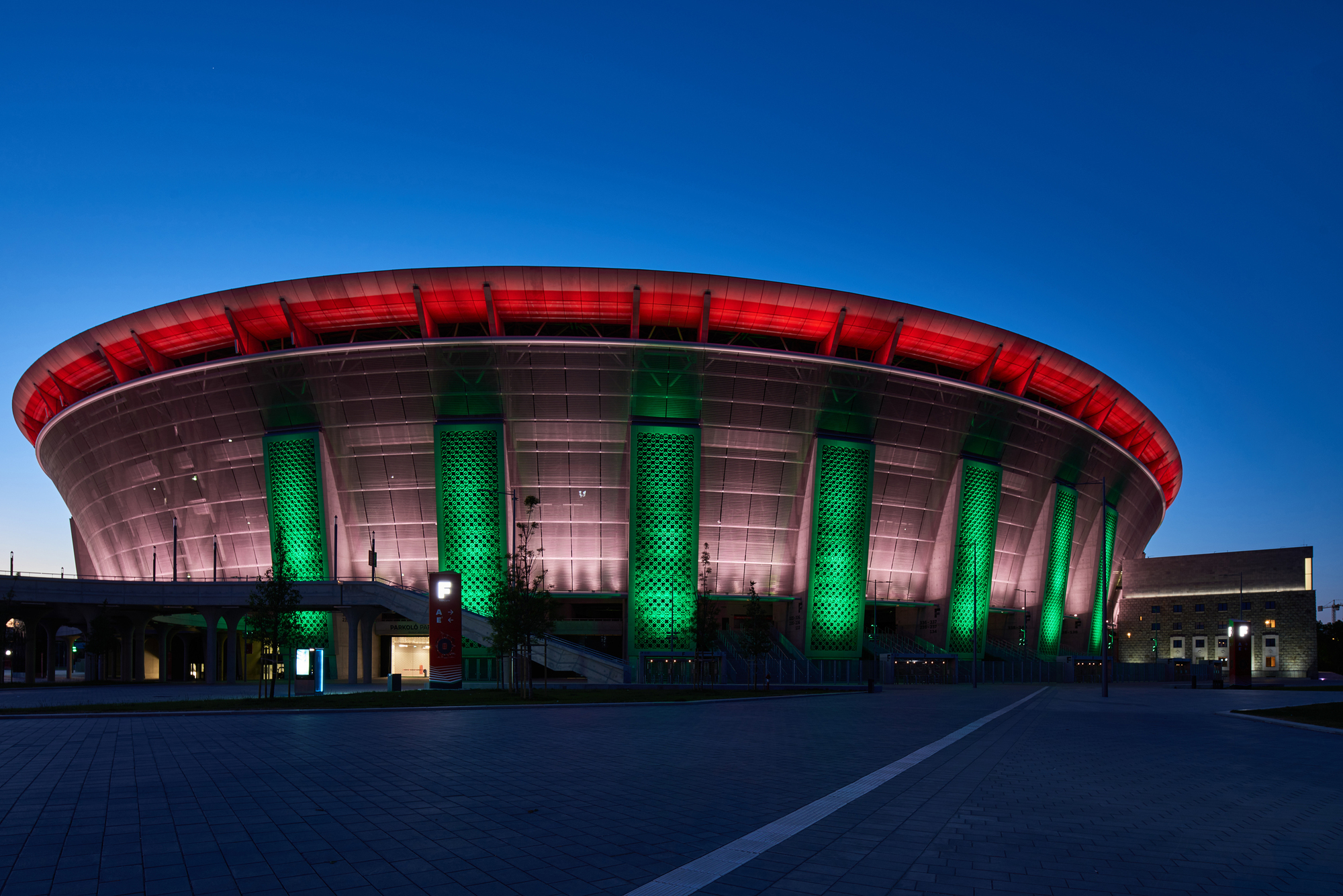 Circular stadium facade with red crown lighting and green illuminated columns at dusk