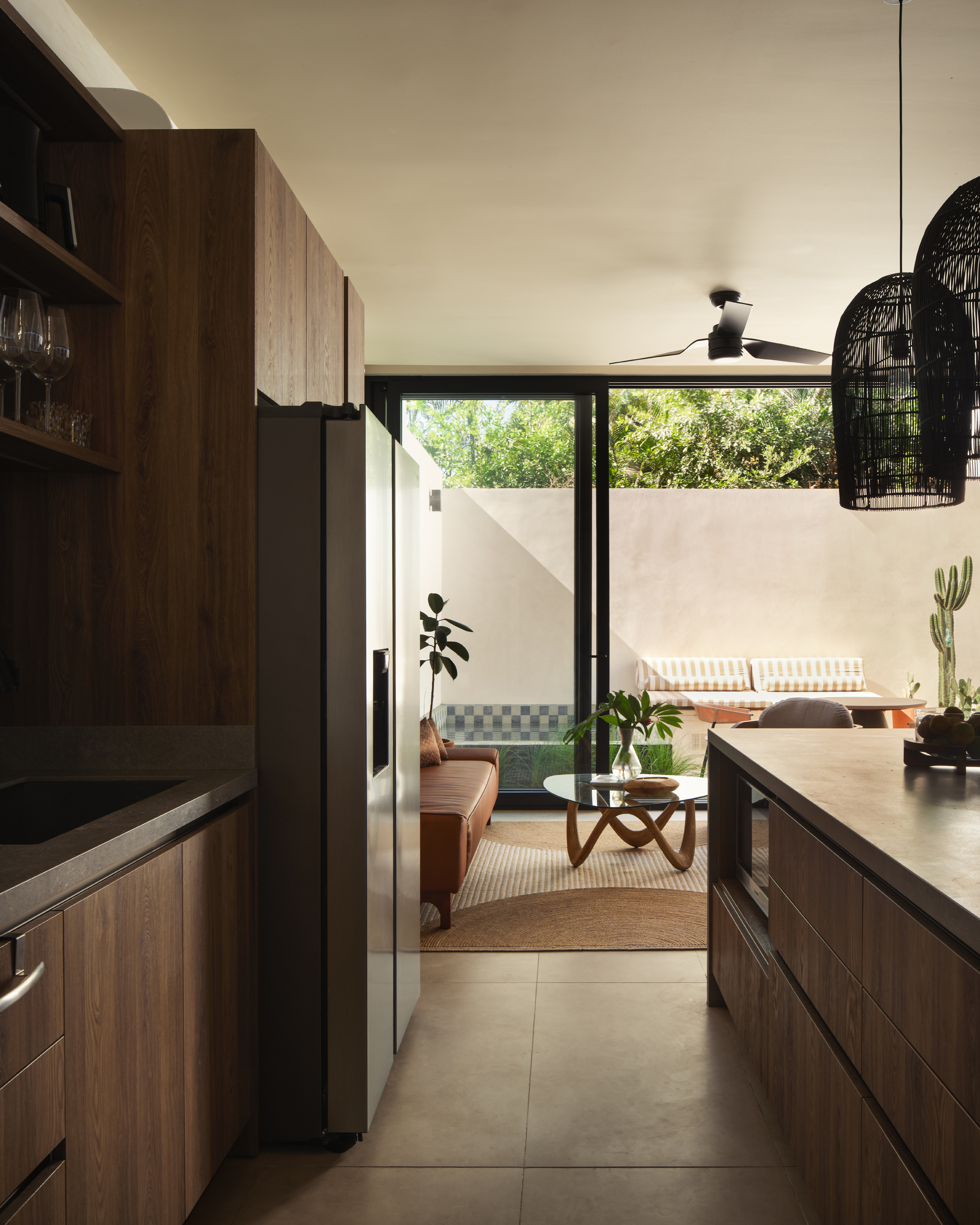 Kitchen with timber cabinetry looking through to a living room and tiled outdoor courtyard in sunlight