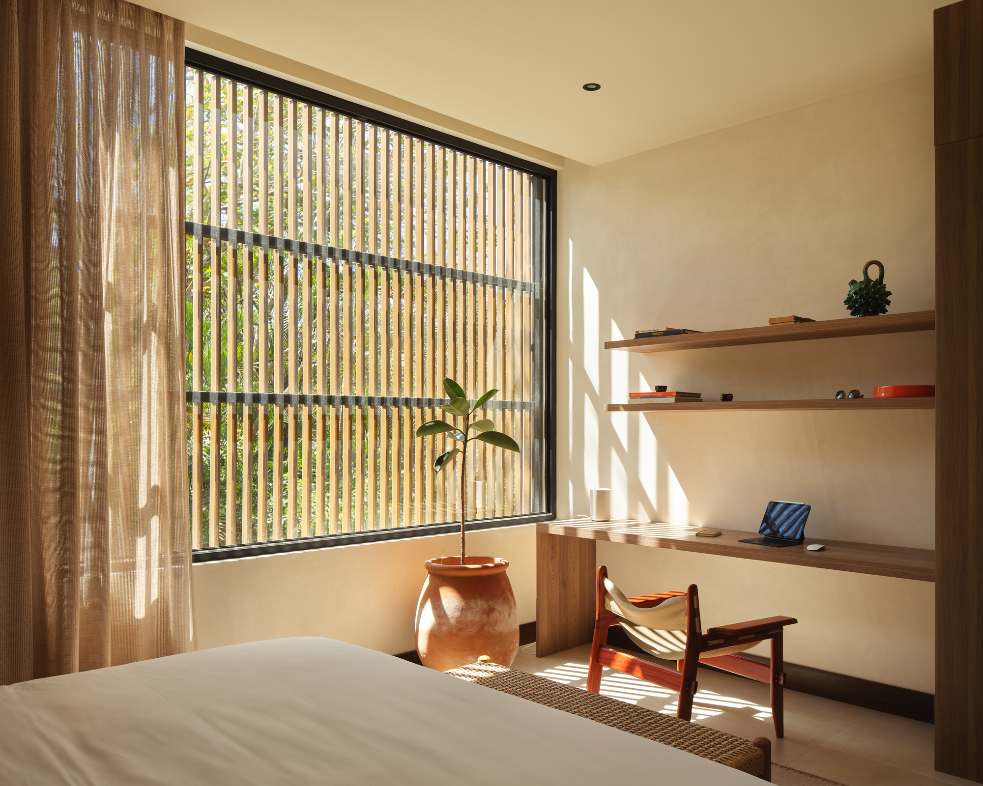 Bedroom with vertical timber screen filtering morning light across walnut shelving and chair
