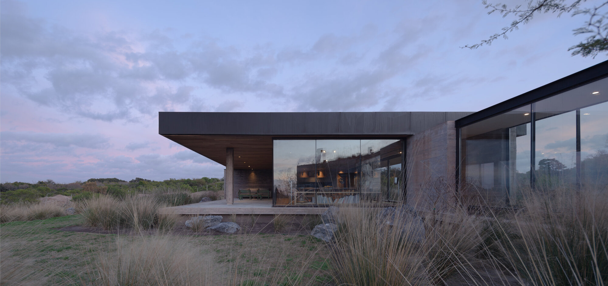 Cantilevered roof sheltering glass-walled living spaces set among grassland at twilight