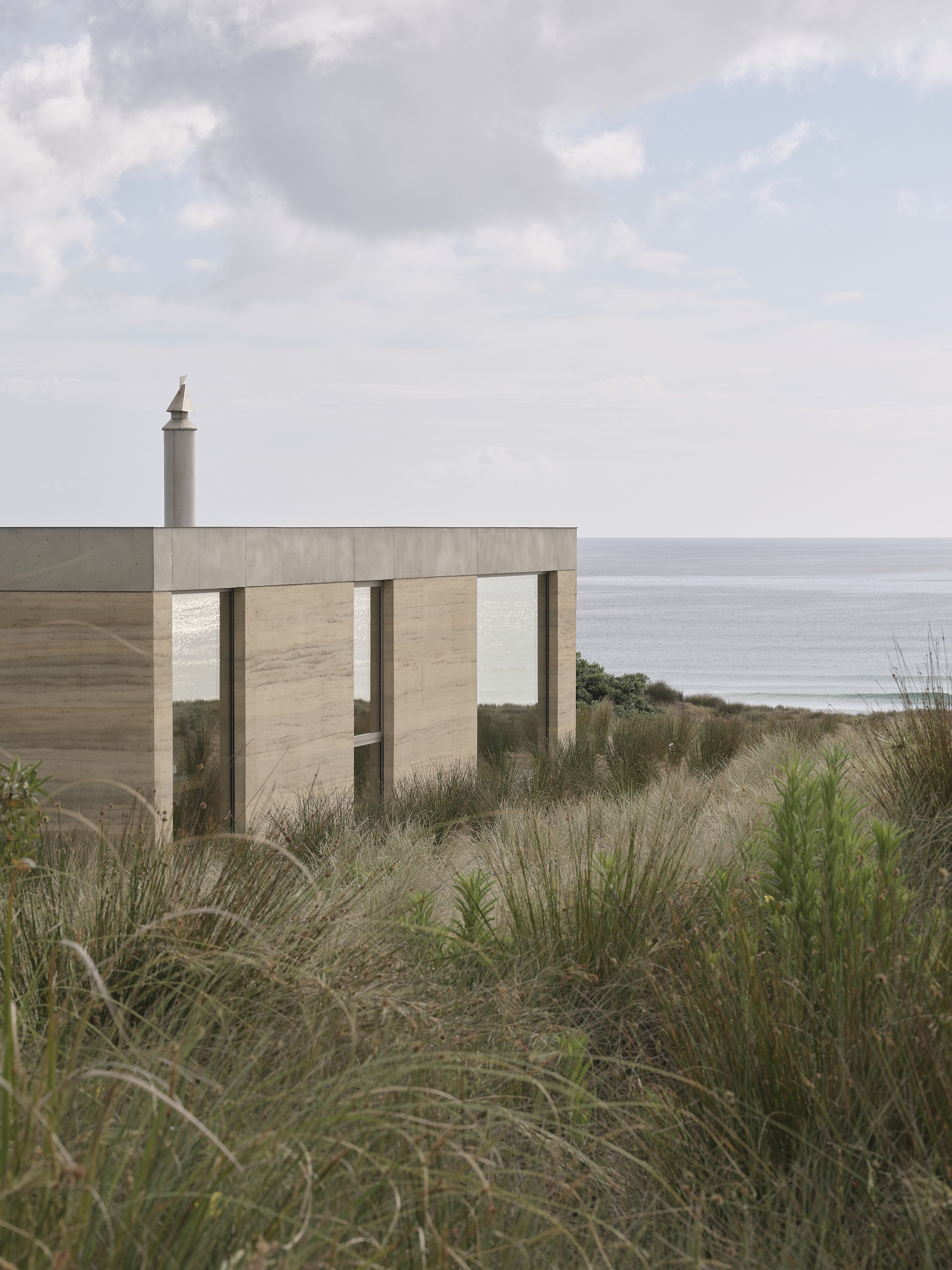 Concrete facade with vertical timber screen openings seen through coastal dune grasses overlooking the ocean