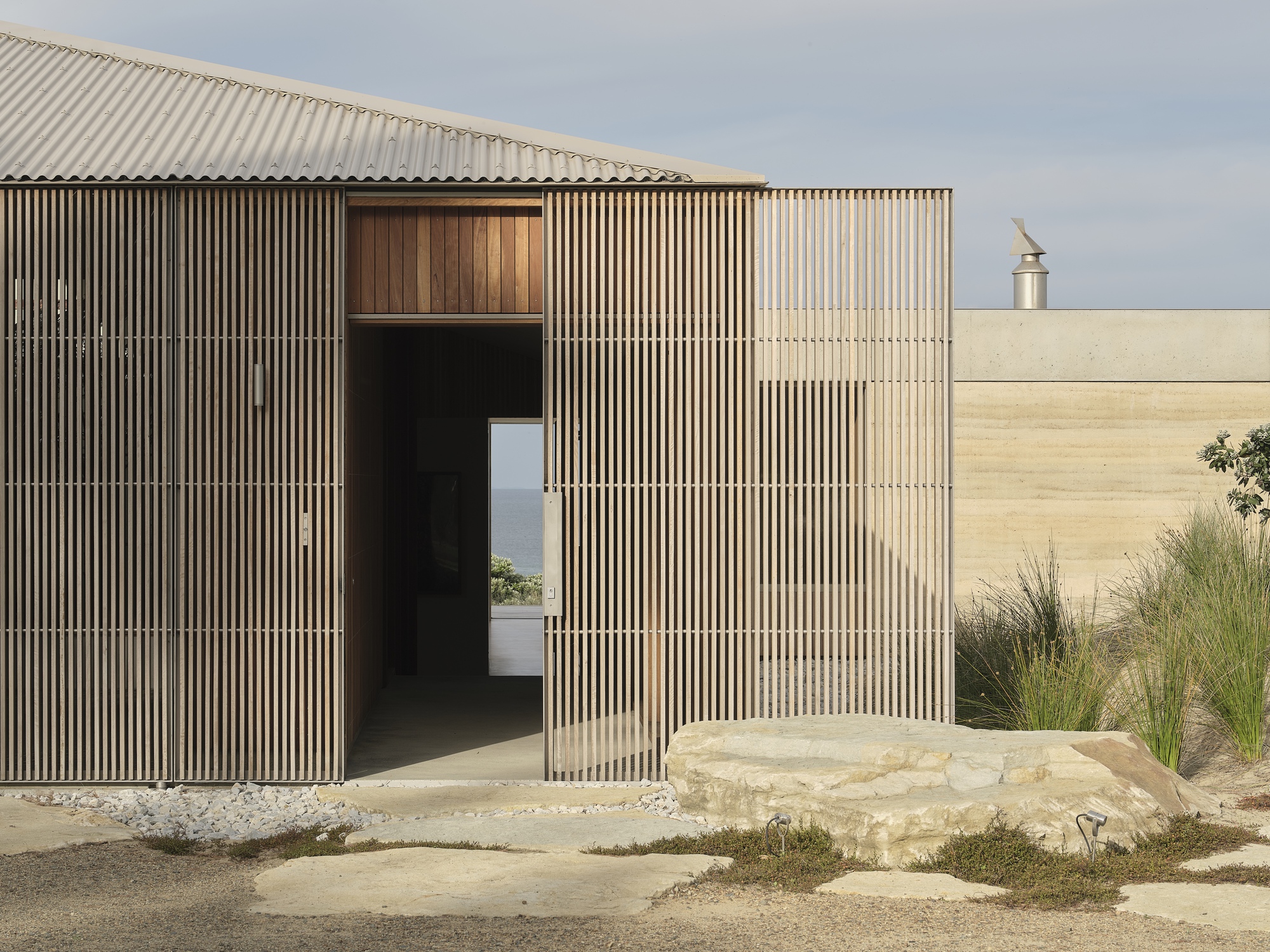 Entry pavilion clad in vertical timber battens with pivoting door and limestone outcrop beside gravel courtyard