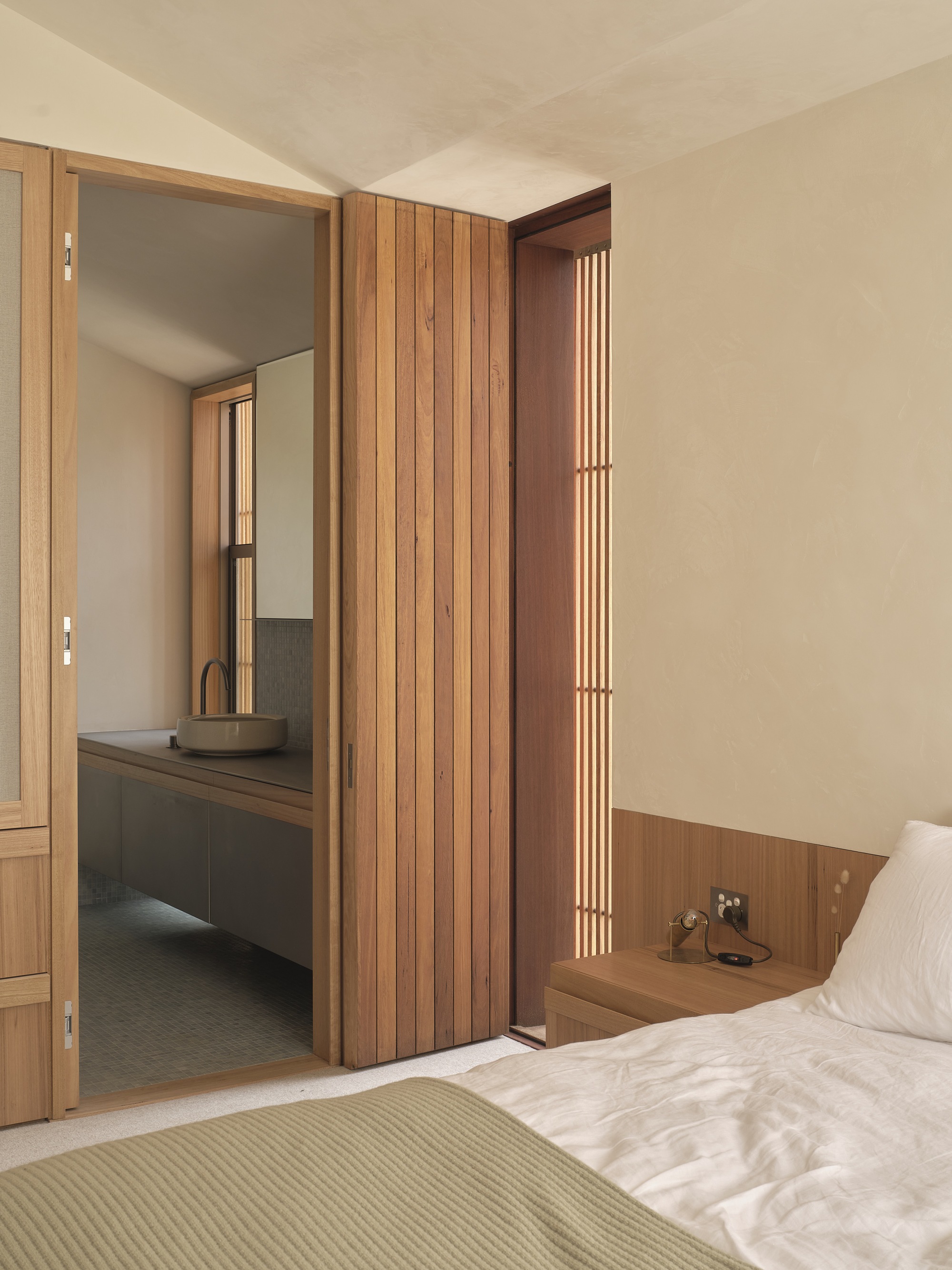 Bedroom view through timber-clad doorway to ensuite bathroom with floating vanity and vertical slatted window