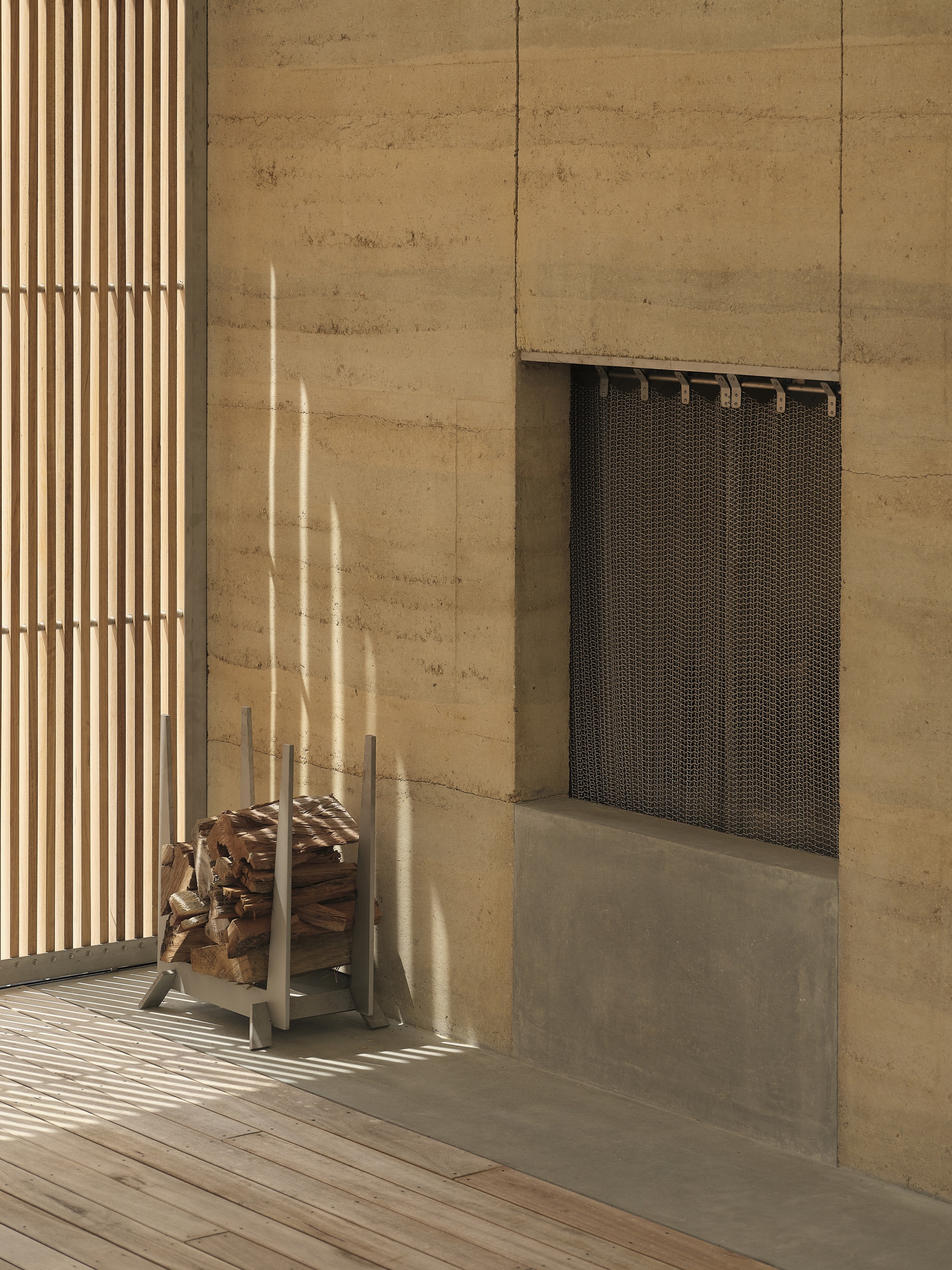 Fireplace recess with metal mesh screen in rammed earth wall beside timber slatted screen in afternoon light