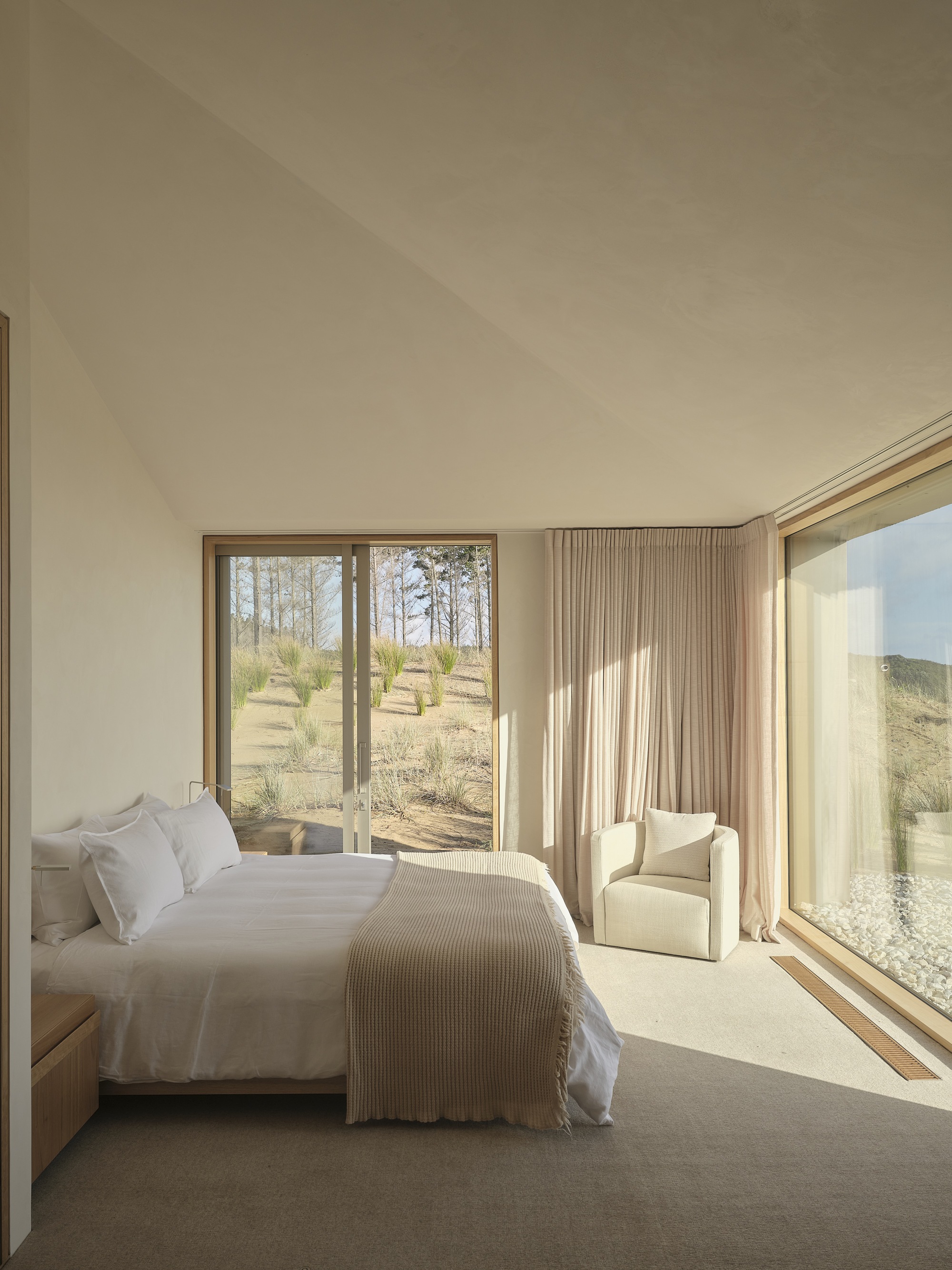 Bedroom with floor-to-ceiling glazing overlooking grassy landscape with scattered trees under soft daylight