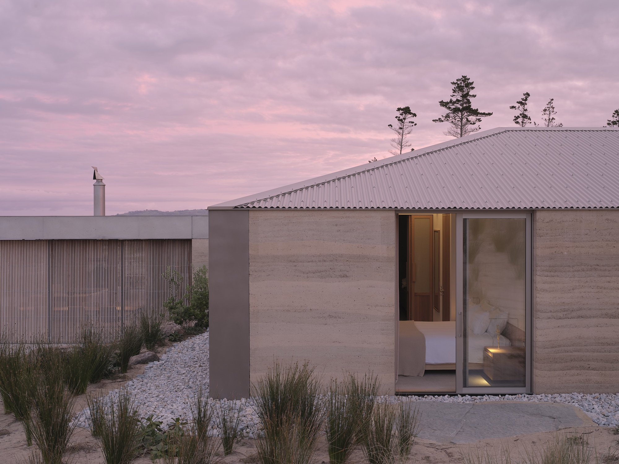 Rammed earth walls and corrugated metal roof among gravel beds and native grasses at dusk