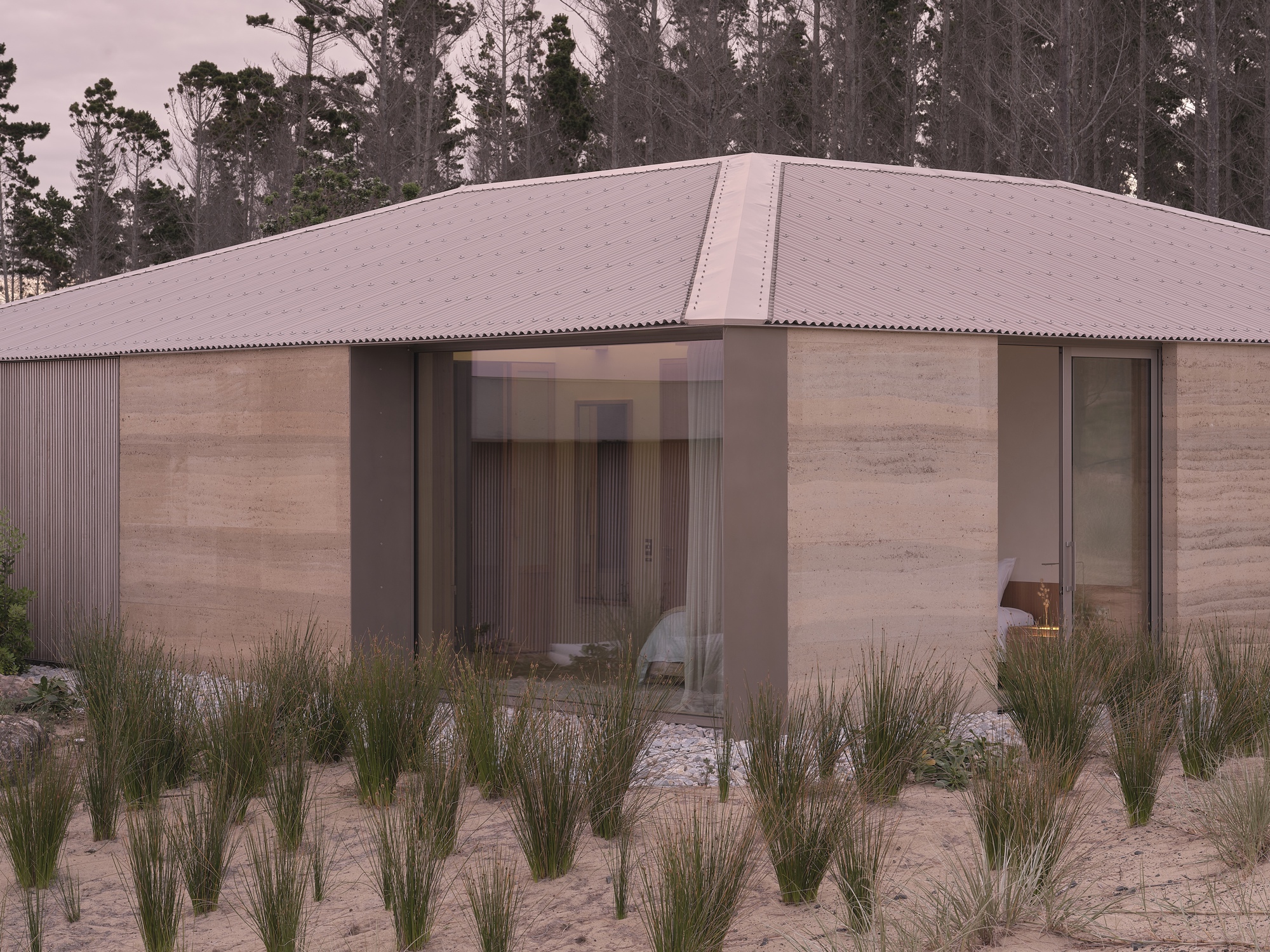 Rammed earth and corrugated metal facade with glazed openings facing sparse grass planting in sandy soil