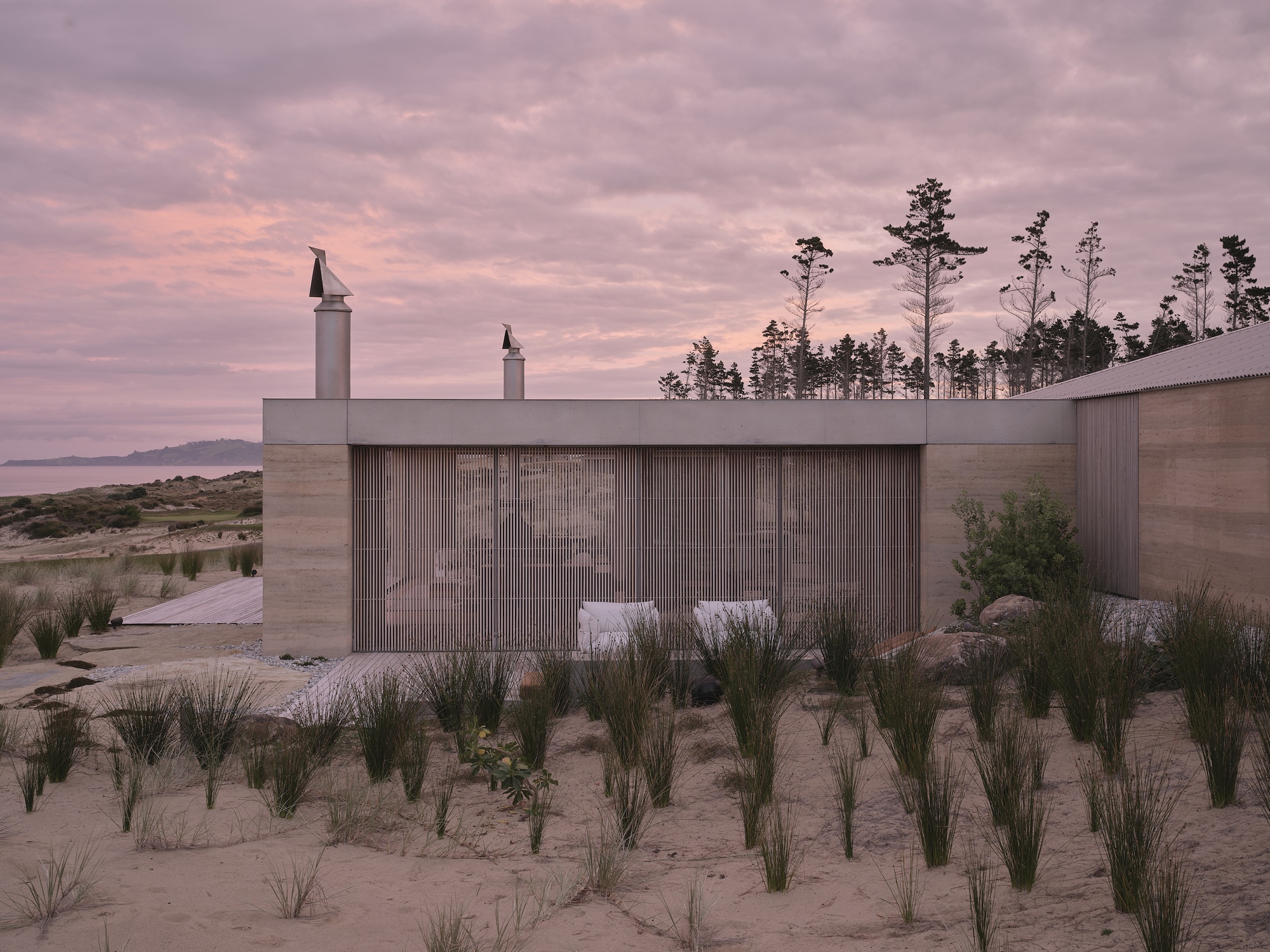 Coastal pavilion with vertical timber cladding and white chimneys surrounded by beach grasses at dusk