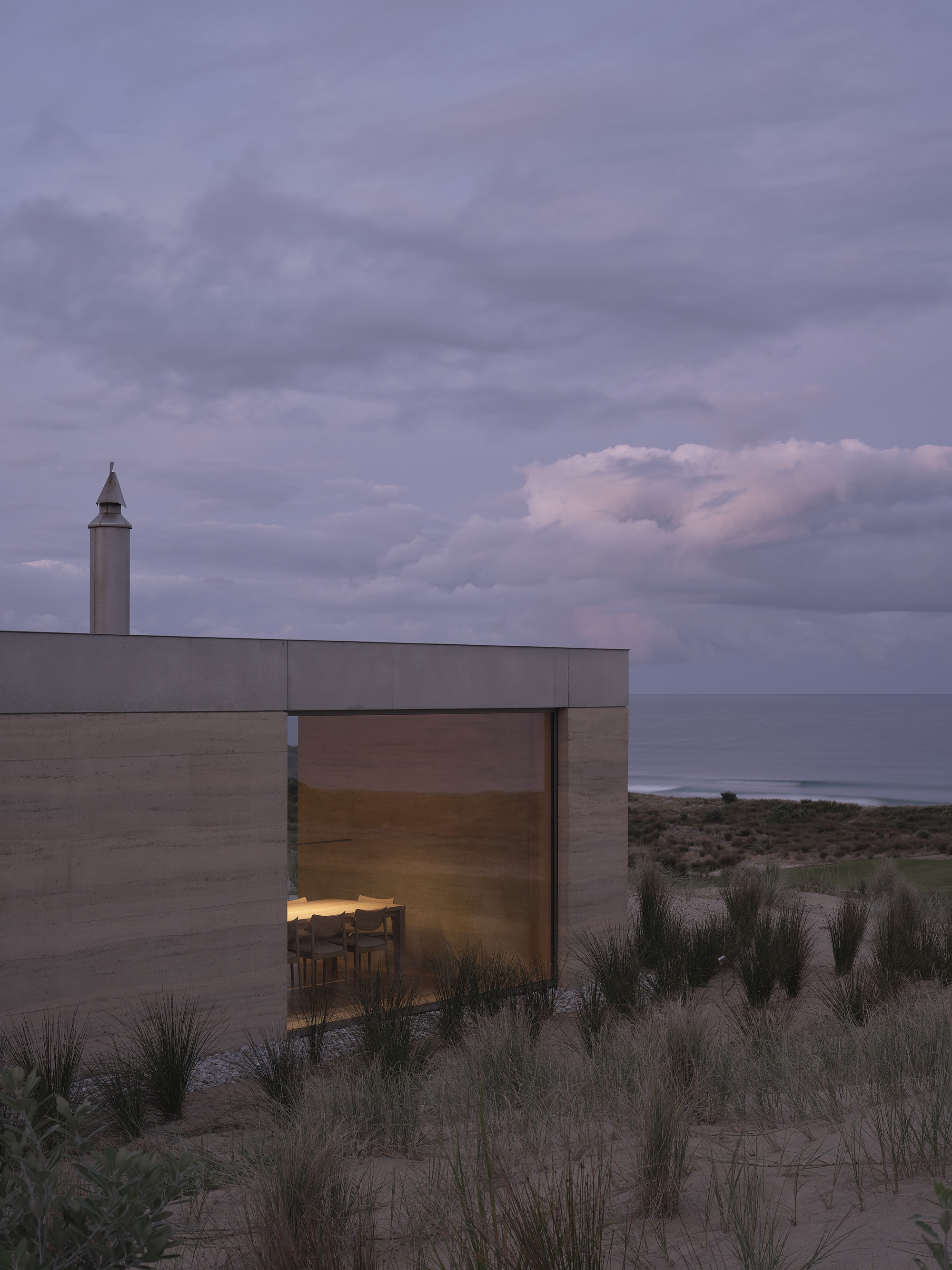 Glass corner room with illuminated interior and coastal view beyond dune grasses at twilight