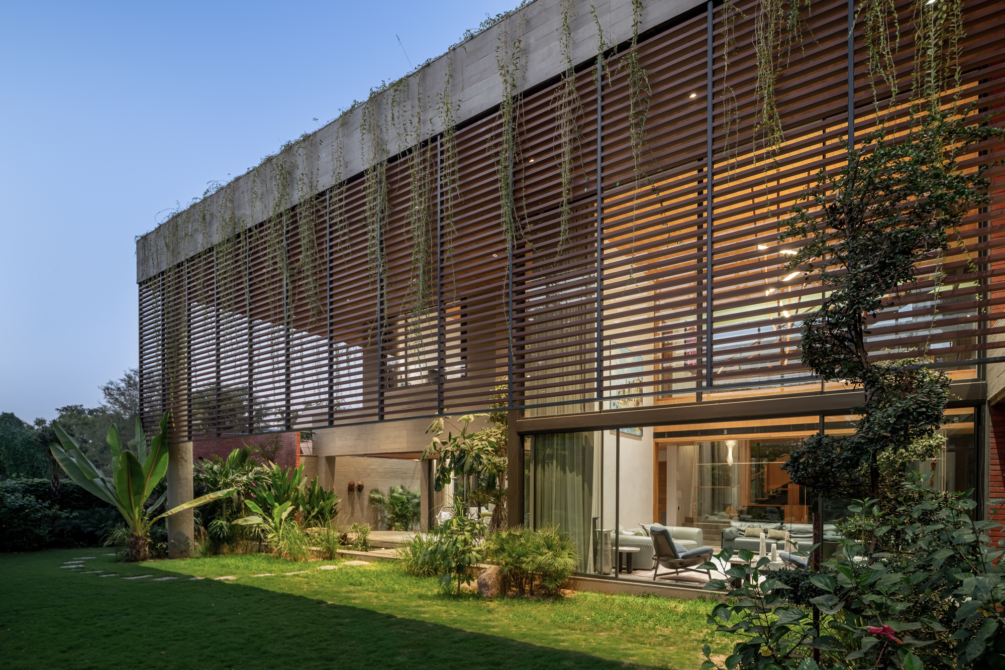 Garden facade at dusk showing illuminated interior behind glass and horizontal timber louvers with cascading plants