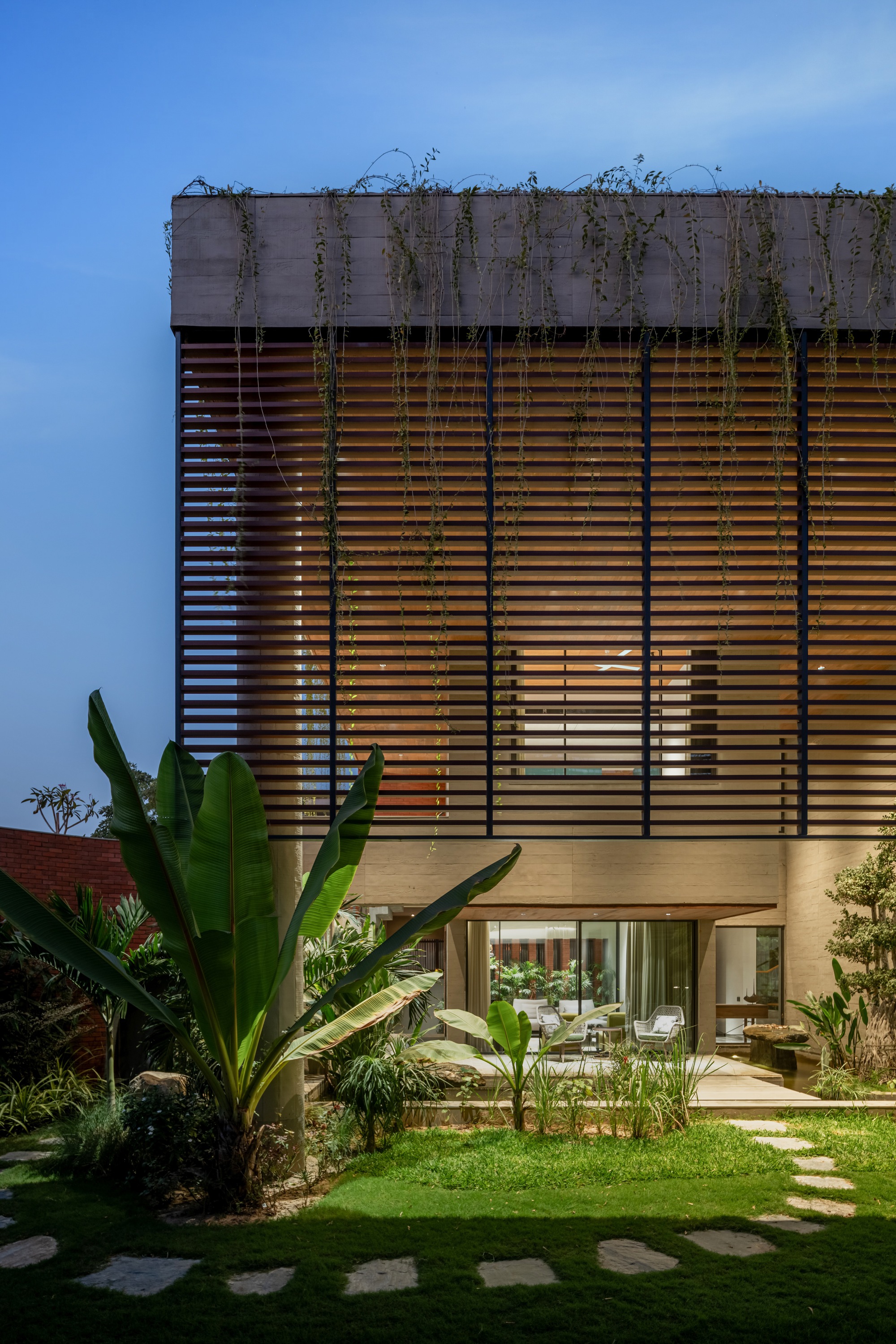 Timber louvered facade beneath a concrete parapet with cascading vines at twilight