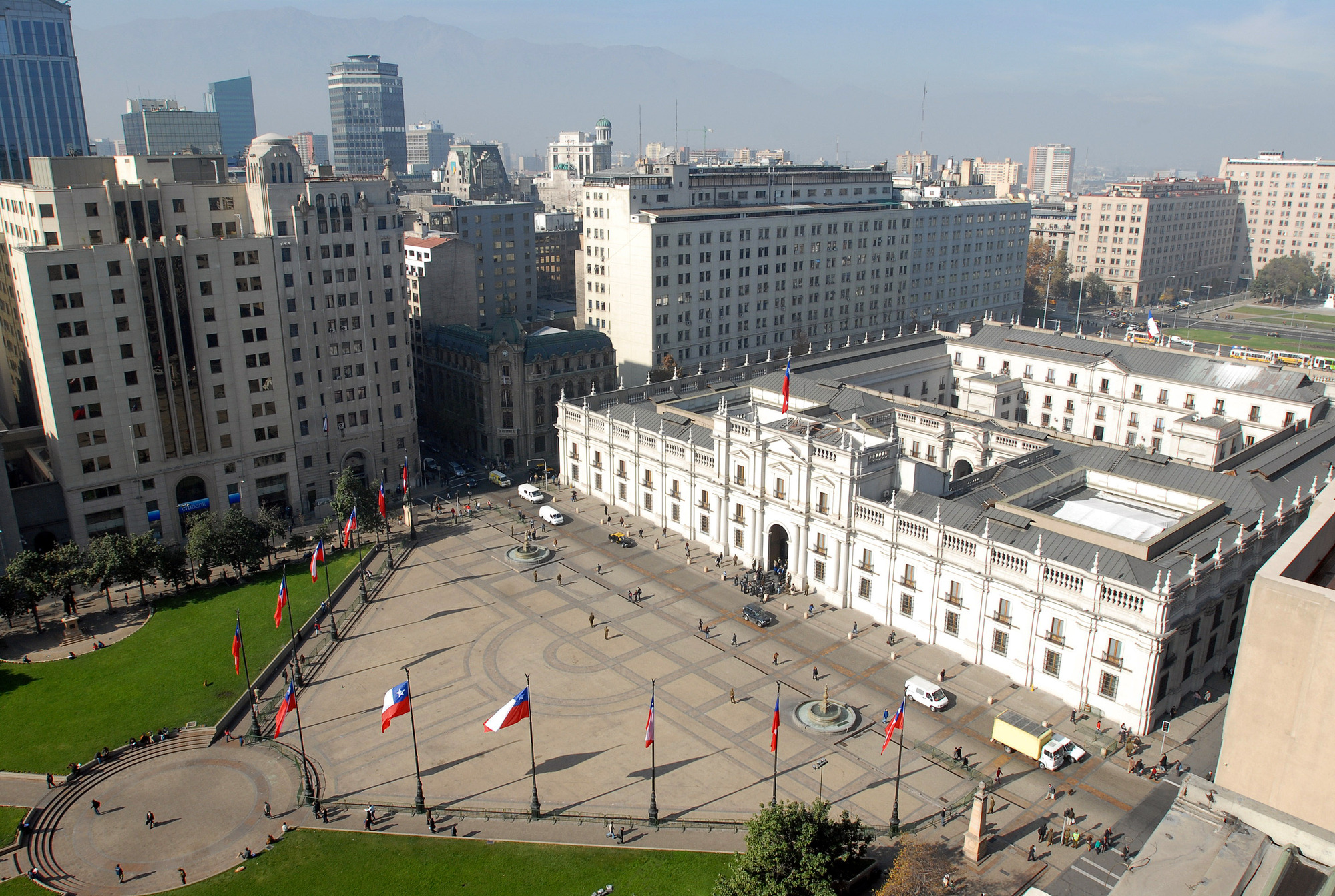 Elevated view of a white palace building with neoclassical facade facing a paved public plaza with flags
