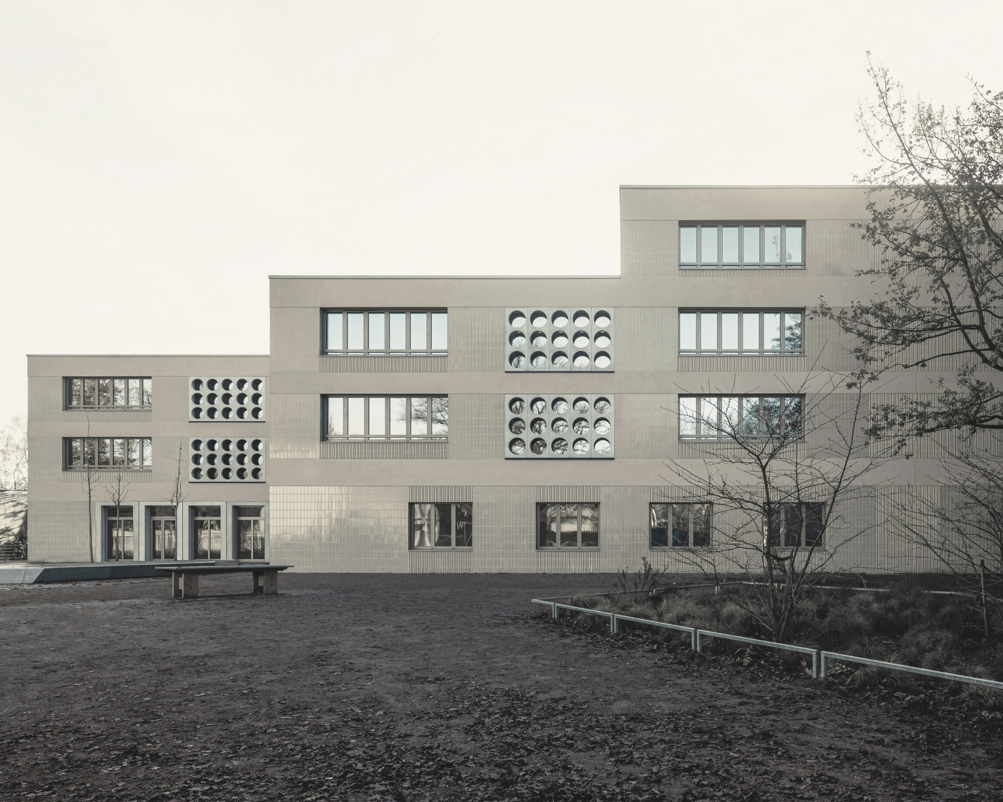 Stepped facade with alternating rectangular windows and circular perforated panels in pale brick under an overcast sky
