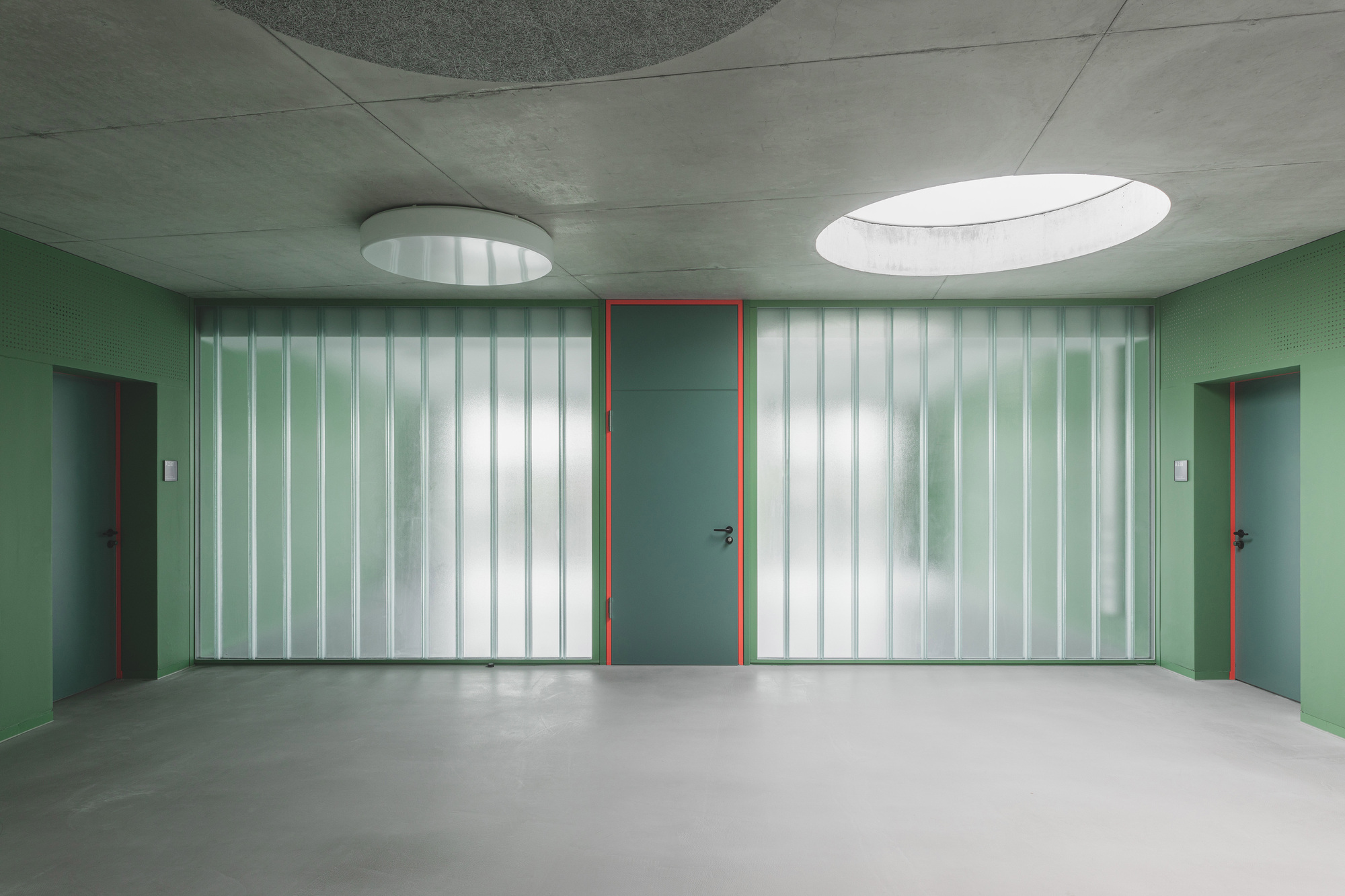 Corridor with translucent vertical glass panels flanking doors painted in green and red under oval skylights
