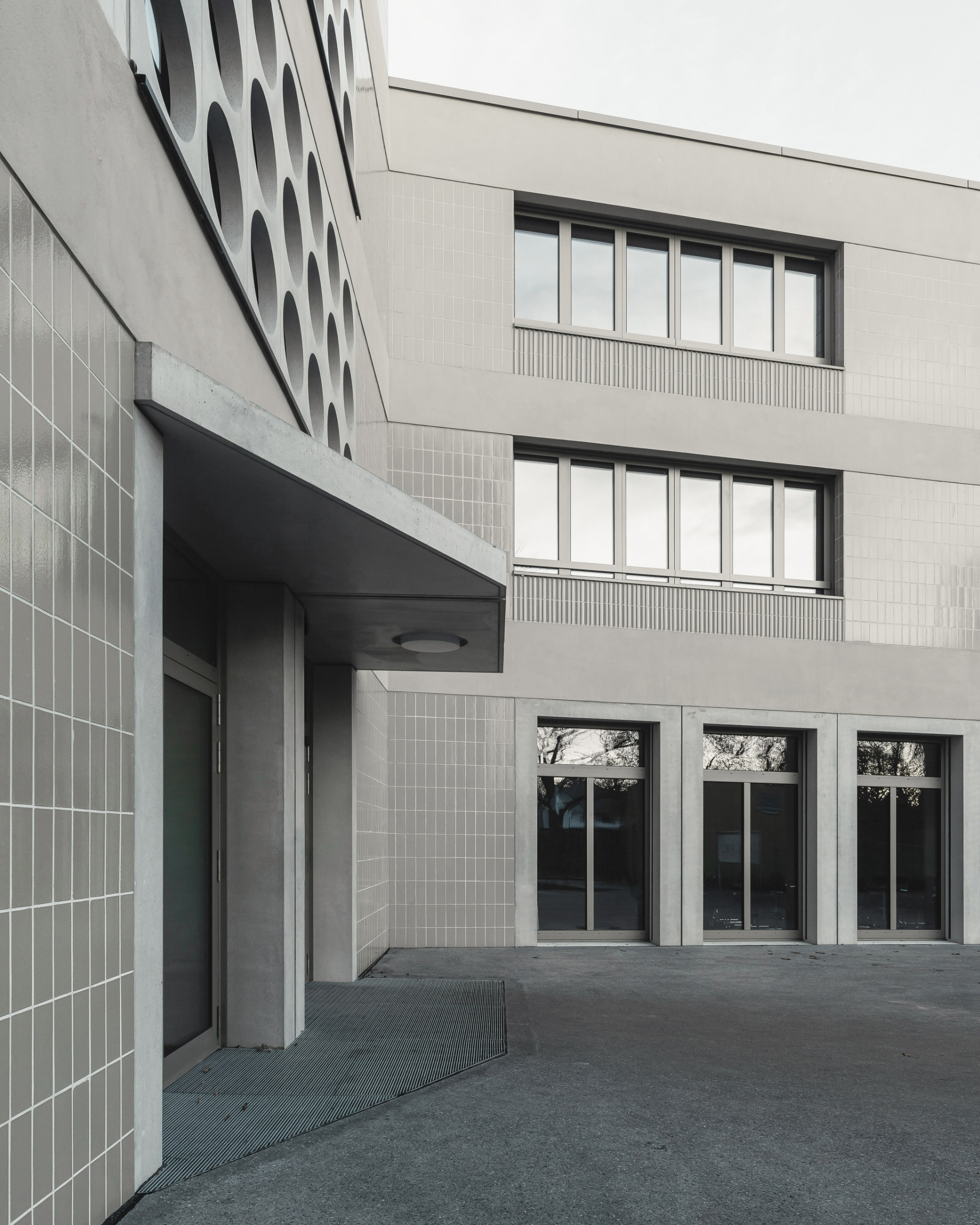 Courtyard entrance with light tile facade, recessed doorway, and horizontal ribbon windows under overcast sky