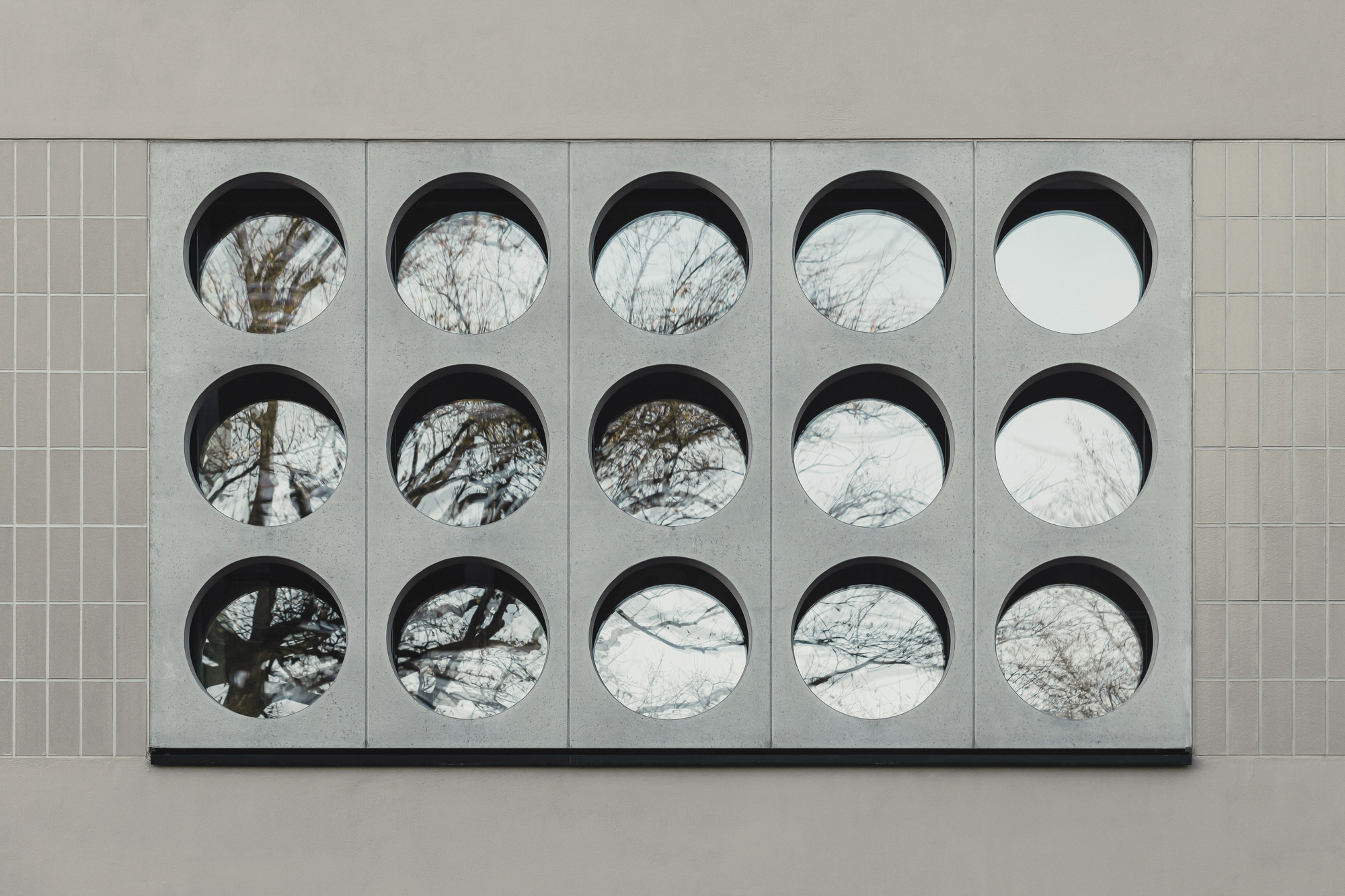 Close-up of fifteen circular openings in concrete panel framing views of bare tree branches