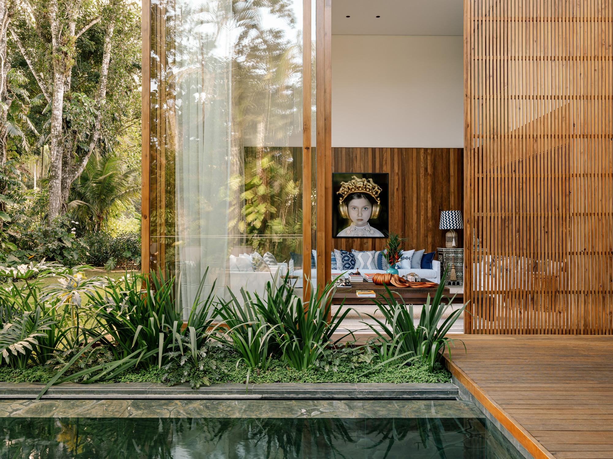 View across reflecting pool with timber deck and planted bed framing a glass-walled living room