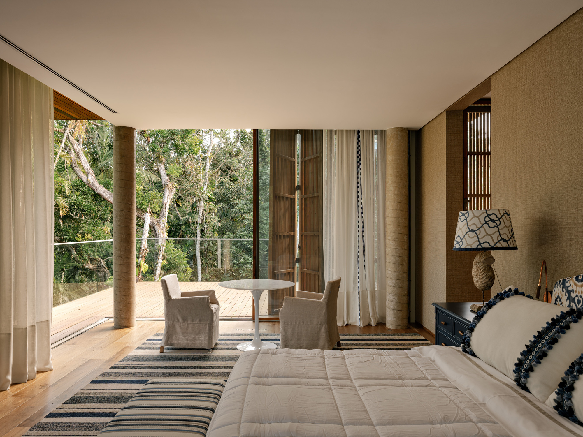 Bedroom opening to timber deck terrace framed by floor-to-ceiling glass and trees beyond