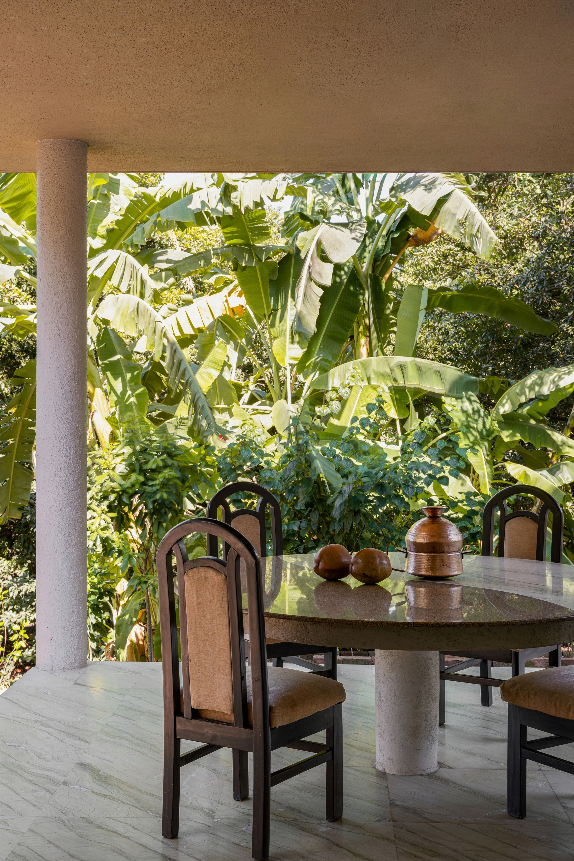 Covered terrace with cylindrical column and dark wood dining chairs facing dense banana plants in bright sunlight