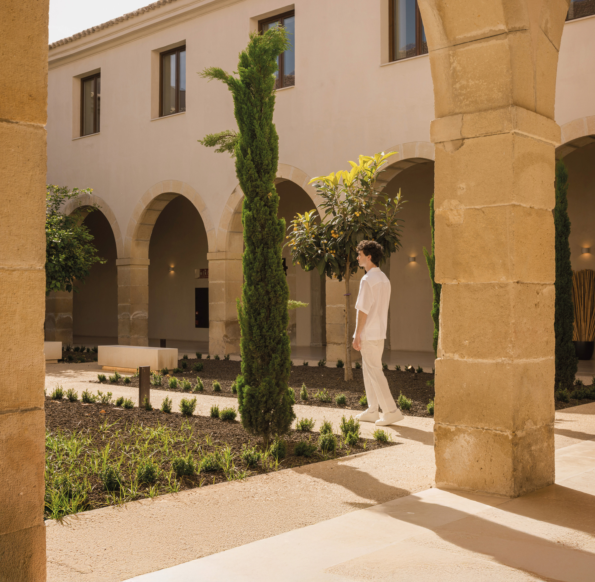 Stone arcade column with cypress trees and planted beds in the courtyard beyond in afternoon light