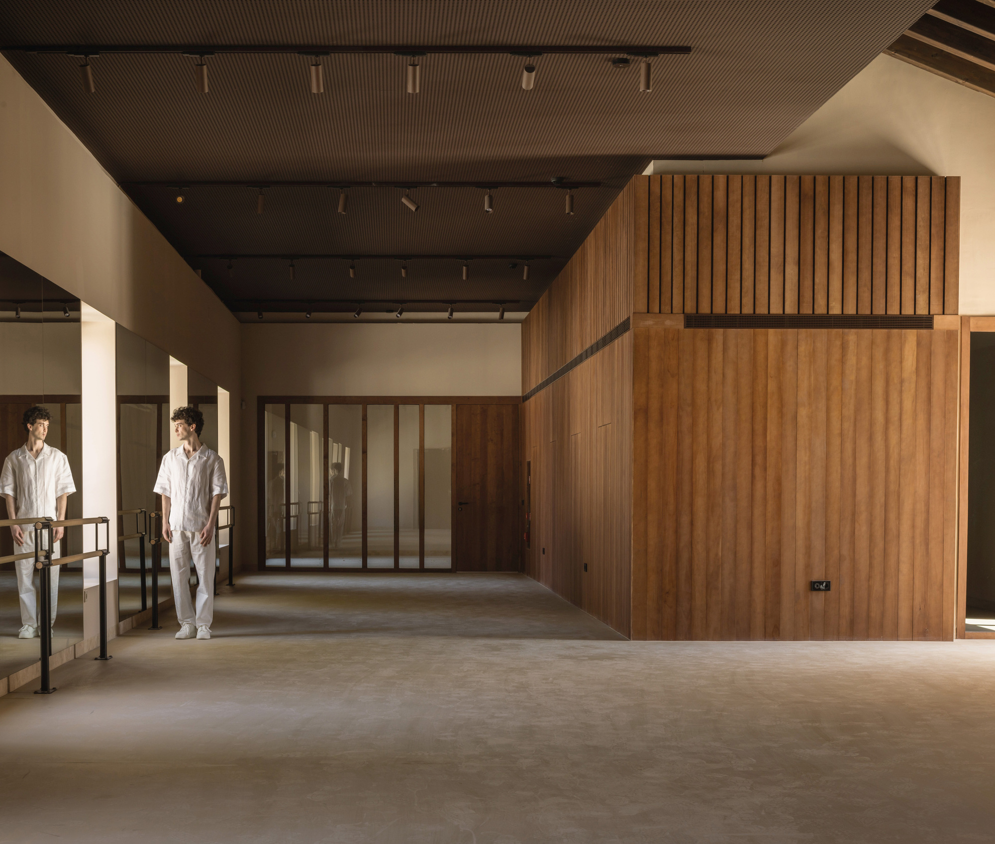 Interior space with timber-clad volume, perforated dark ceiling panel, and two figures near the railing