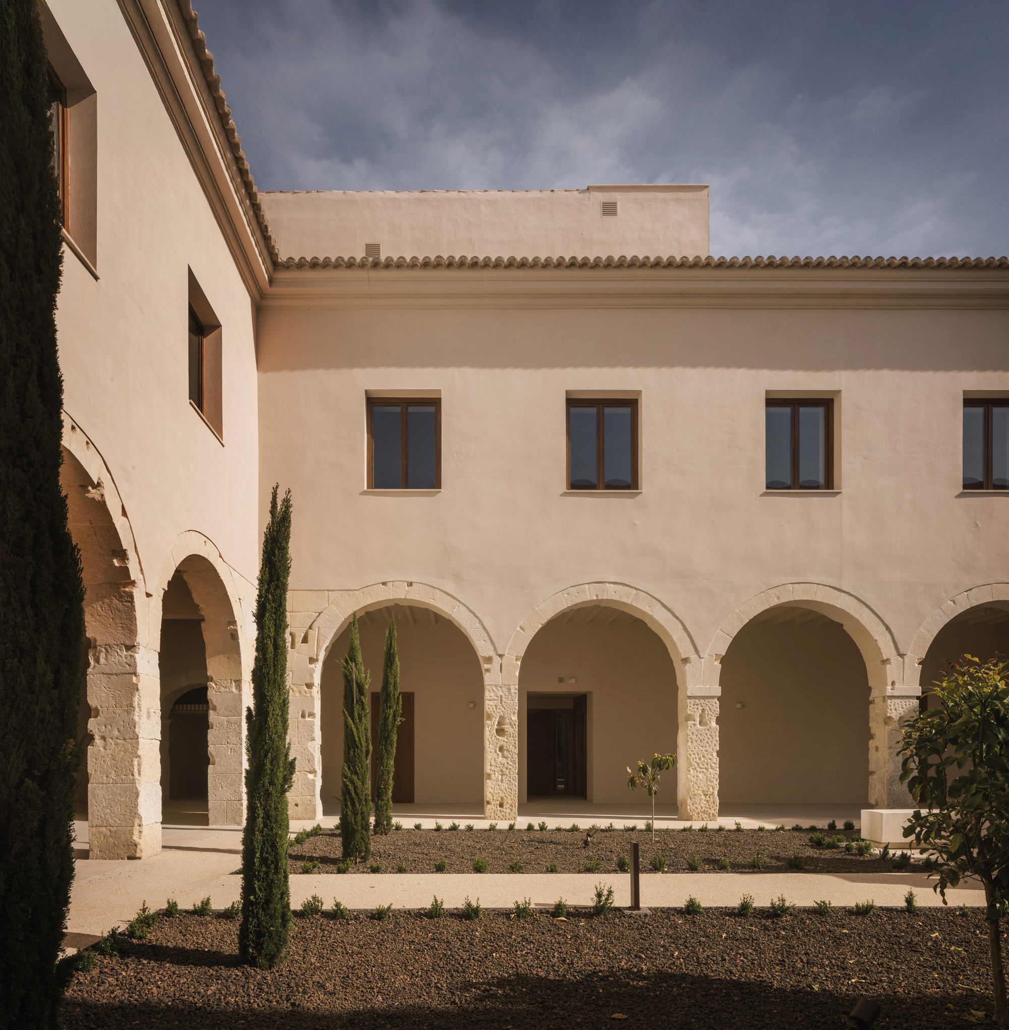 Courtyard facade with stone arcades, plastered upper level, cypress trees, and gravel planted beds below