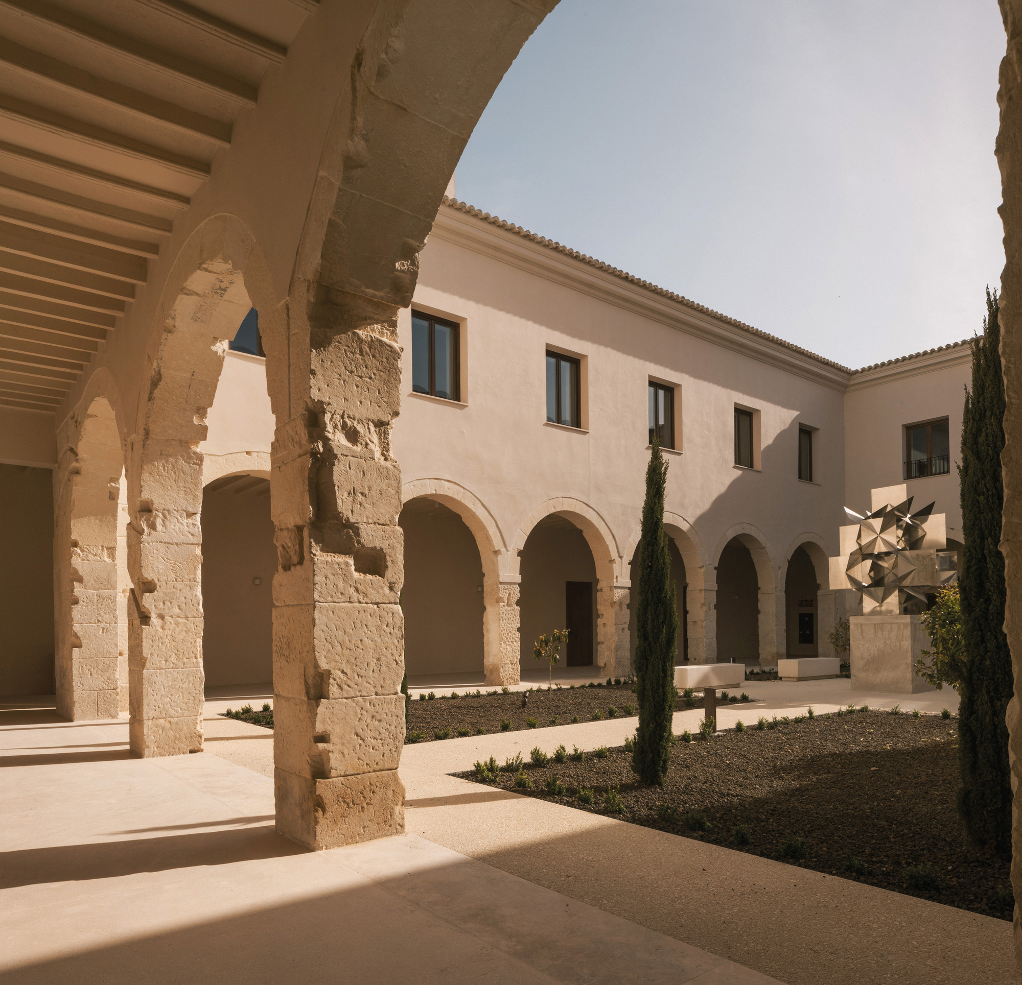 Courtyard view through restored stone arches with cypress trees and planted beds in dappled sunlight